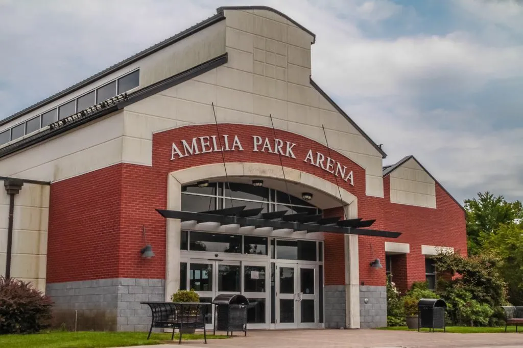 La entrada principal de ladrillo de la arena bajo un frontón arqueado con las letras "Amelia Park Arena", con puertas de vidrio y una banca al frente. — Amelia Park Arena