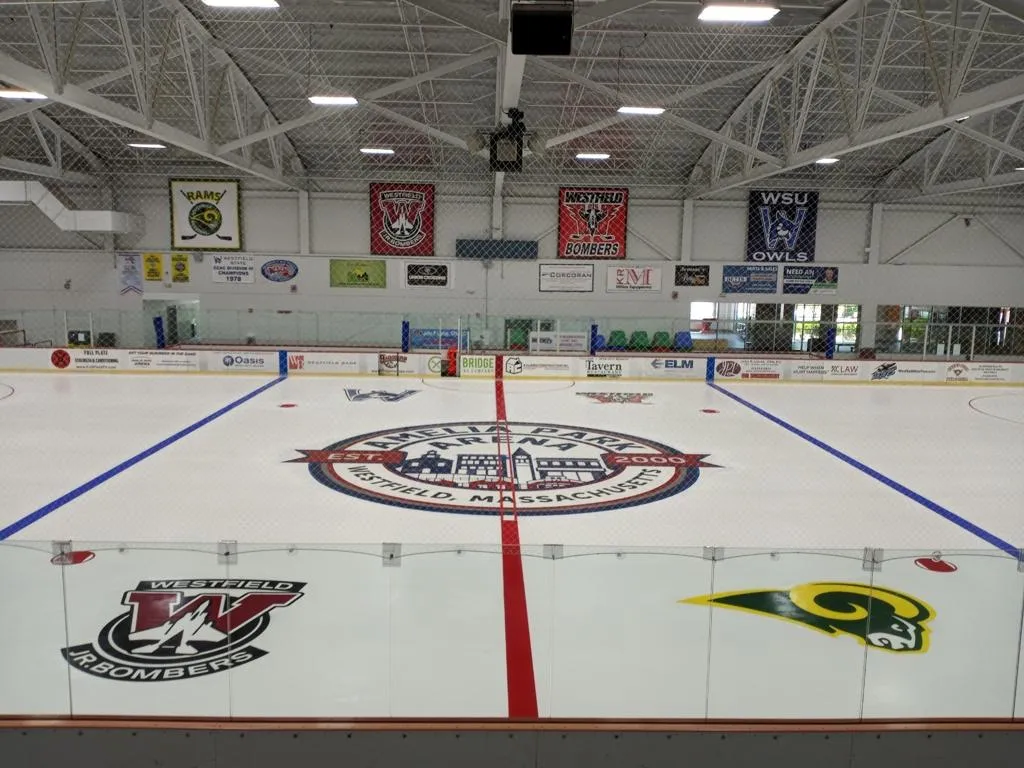 La pista vacía desde detrás del vidrio en el centro del hielo: el escudo de Amelia Park al centro, los logotipos de los Westfield Bombers y de Westfield State en el hielo y mantas de equipos a lo largo de la pared del fondo. — Amelia Park Arena