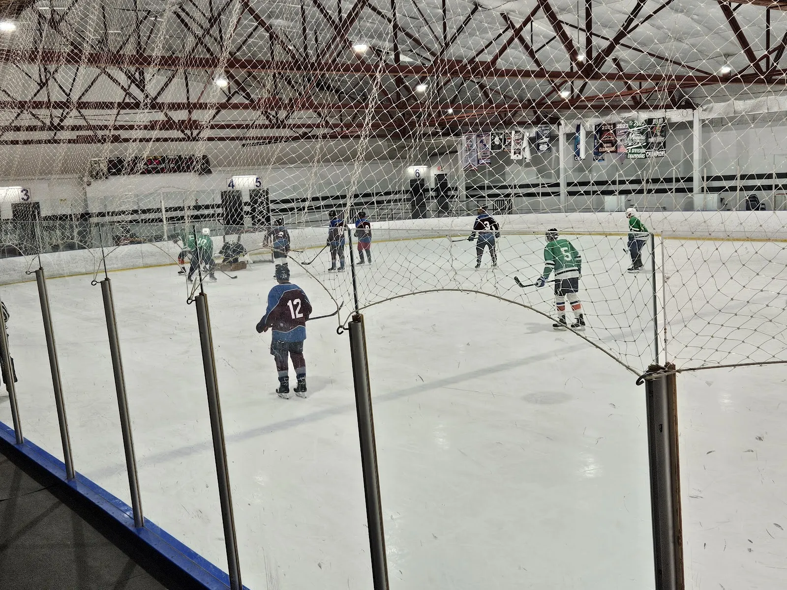 A game seen from the glass at one end, players spread across a white sheet under a red-trussed roof, with spectators' netting along the boards. — Ameri-Can North Sports Center