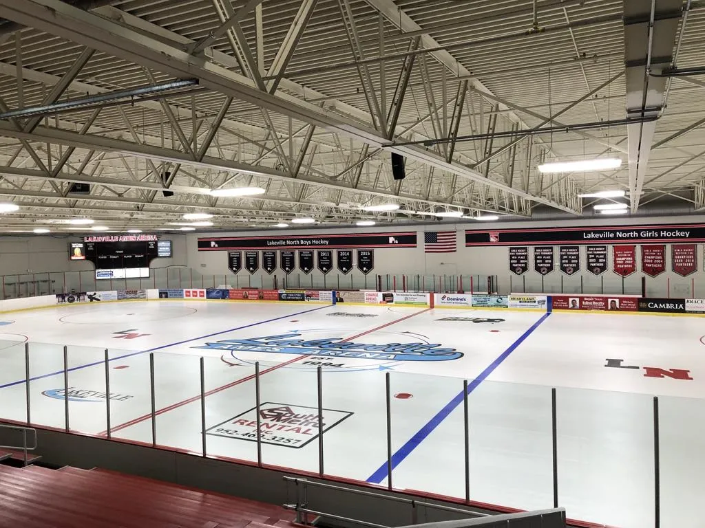 Pista vacía de tamaño NHL vista desde las gradas, con un logotipo de "Lakeville Ames Arena" pintado en el centro de la pista, un marcador con el nombre de la arena y mantas de campeonato de hockey en la pared. — Lakeville Ames Arena (Lakeville Arenas)
