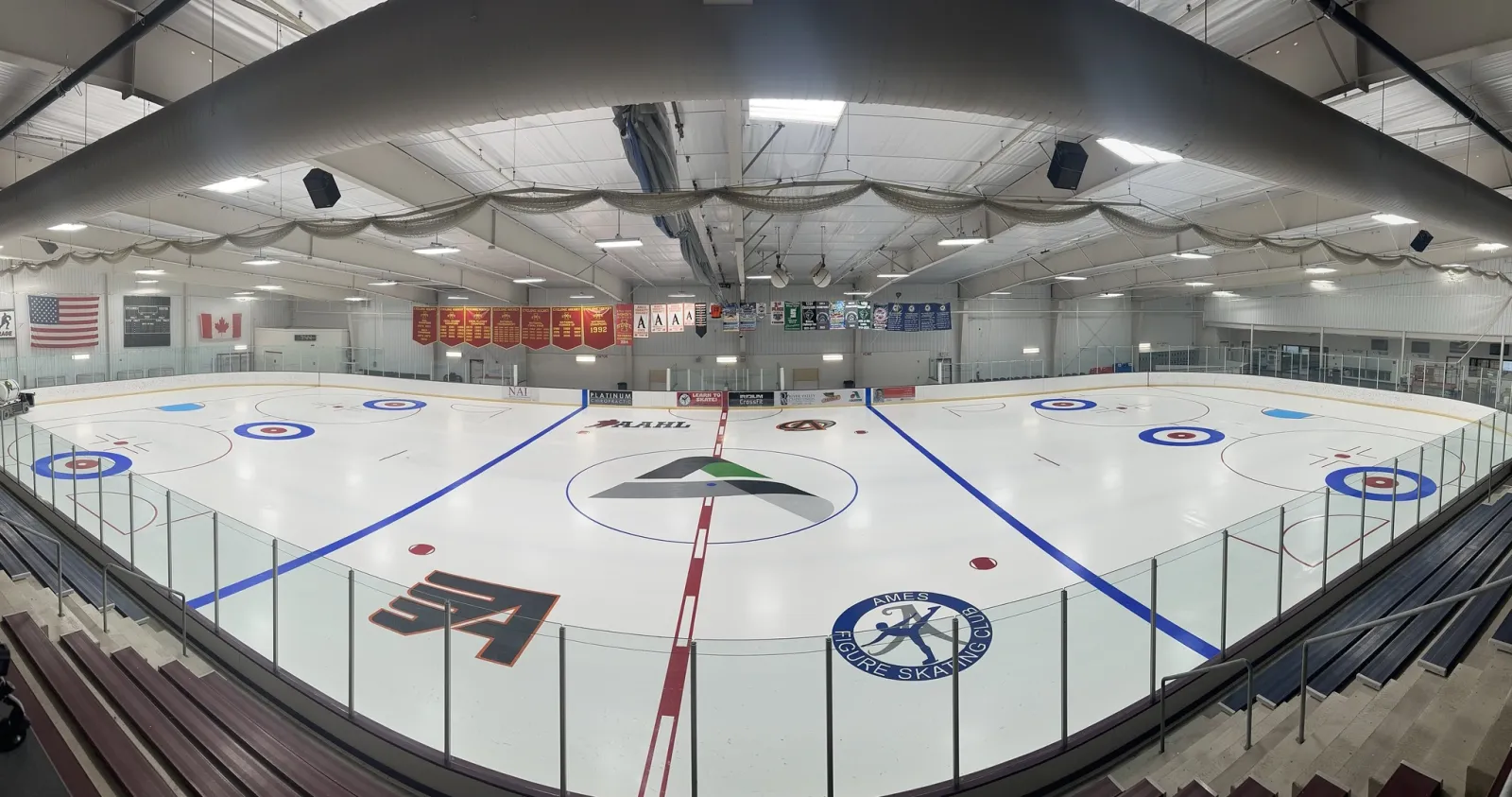 Wide panoramic view of the full ice sheet from the stands, showing the Iowa State "ISU-A" logo and Ames Figure Skating Club logo painted on the ice, championship banners on the far wall, and bleacher seating in the foreground. — Ames/ISU Ice Arena