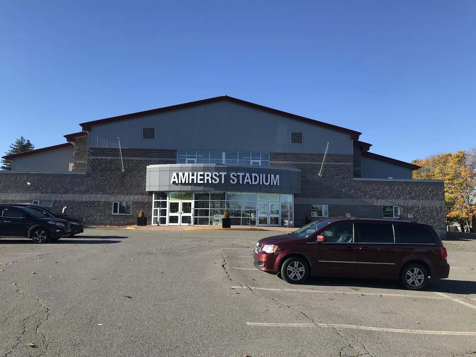 La fachada del Amherst Stadium, un pabellón gris con revestimiento de piedra y su nombre sobre una entrada acristalada, visto al otro lado del aparcamiento con dos vehículos estacionados bajo un cielo azul despejado. — Amherst Stadium