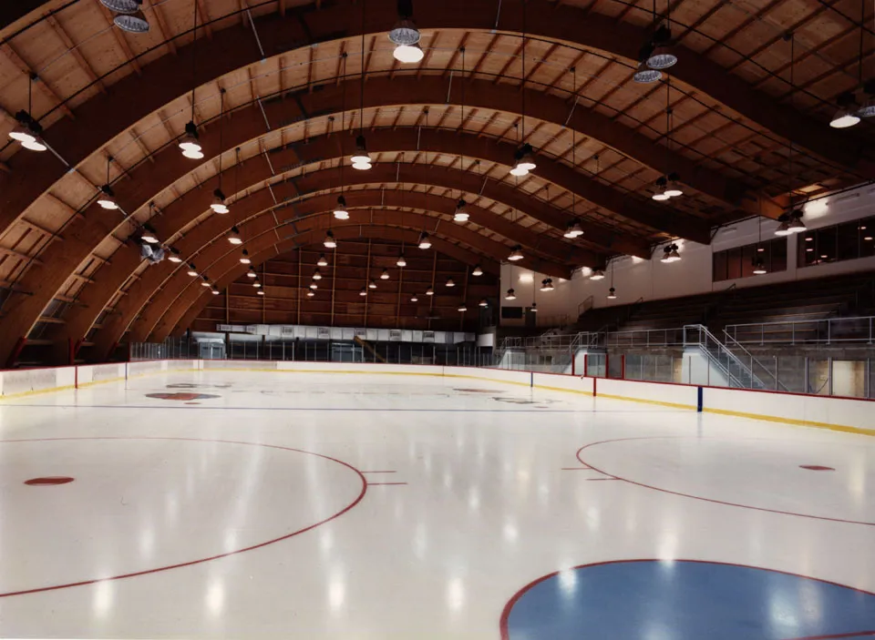 Pista de hielo vacía vista a nivel de la pista bajo el techo curvo del edificio con vigas de madera laminada a la vista, con gradas escalonadas a un lado — Anaheim ICE
