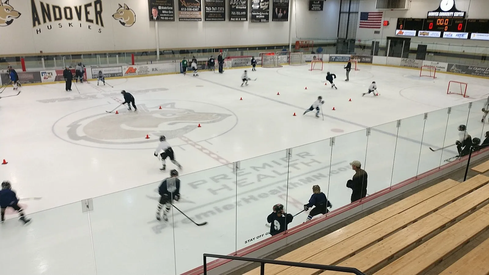 A game seen from the wooden bench along the glass, players spread across a wide sheet under a high roof with a large Andover sign on the far wall. — Andover Community Center