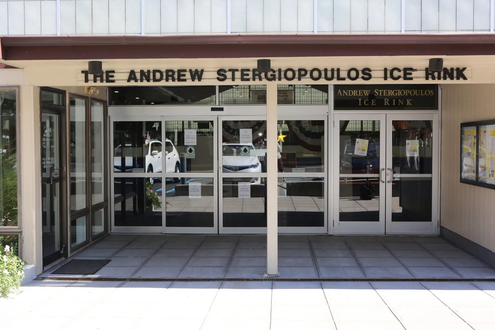 Front entrance doors with 'THE ANDREW STERGIOPOULOS ICE RINK' lettering above the doors and a matching 'Andrew Stergiopoulos Ice Rink' sign to the right. — Andrew Stergiopoulos Ice Rink