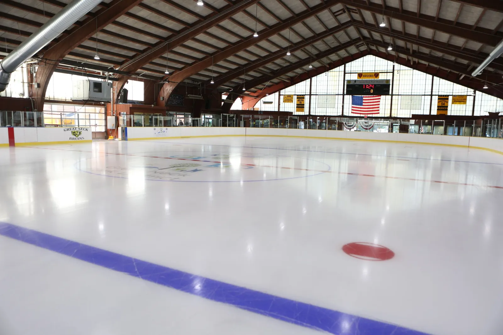 Empty ice sheet under the arena's arched wood-truss roof, with 'GREAT NECK BRUINS' painted on the boards and championship banners hanging above the far end. — Andrew Stergiopoulos Ice Rink