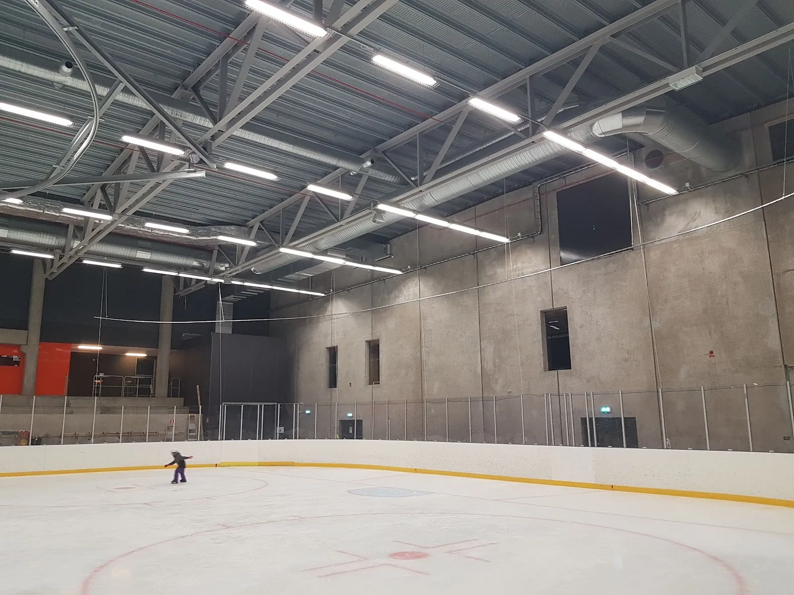 Inside the ice hall of Angered Arena, with the concrete wall and steel roof trusses behind the boards and a single skater at the far left — Angered Arenas ishall