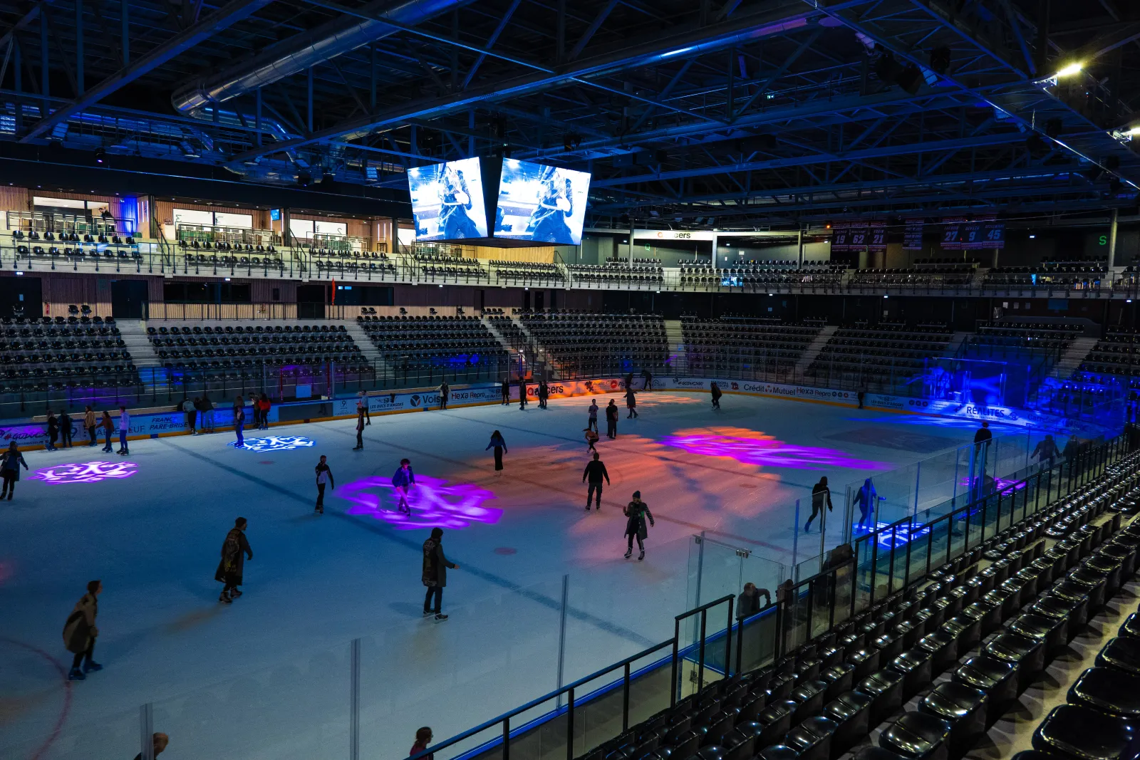 The arena seen from the stands during an evening public session, with coloured light projected on the ice, rows of seats around the sheet and skaters spread across it. — Angers ICEPARC