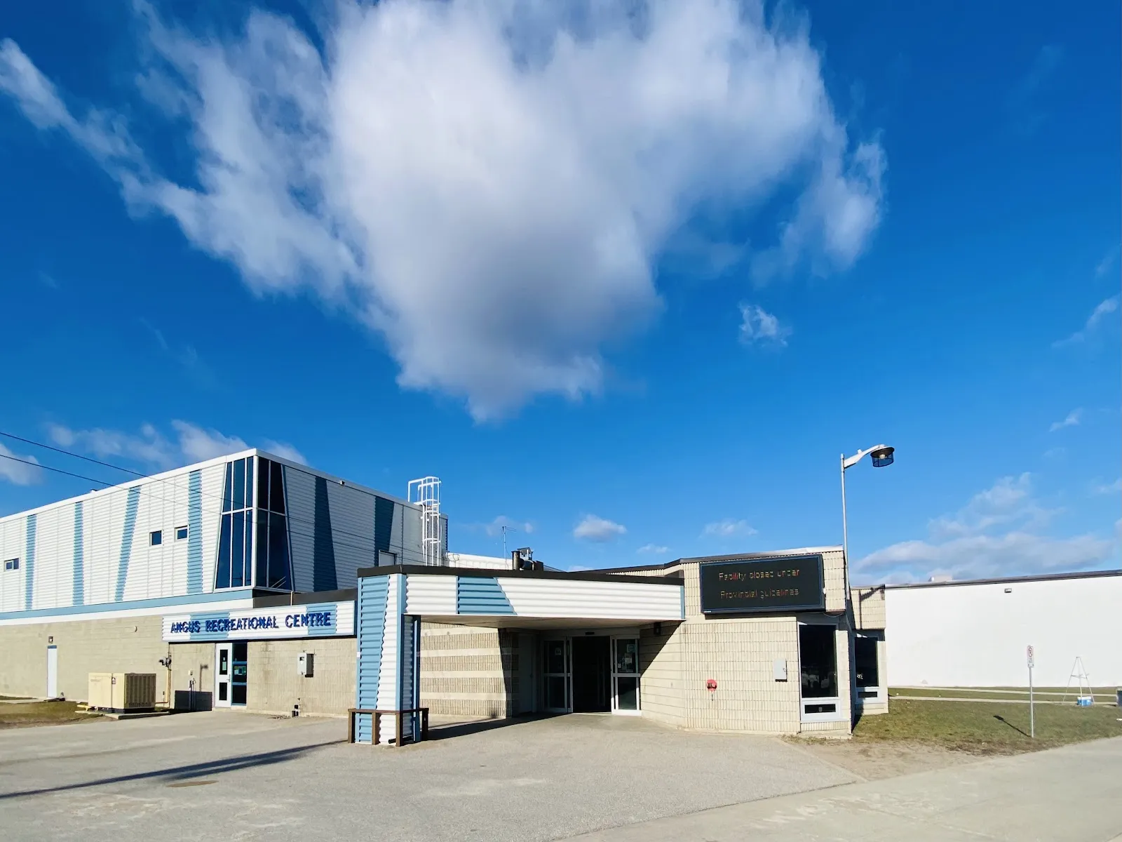 The entrance of the Angus Recreational Centre under a blue sky: the covered doorway, the name sign on the beige brick wall and the blue-and-white striped arena block rising behind it. — Angus Recreation Centre