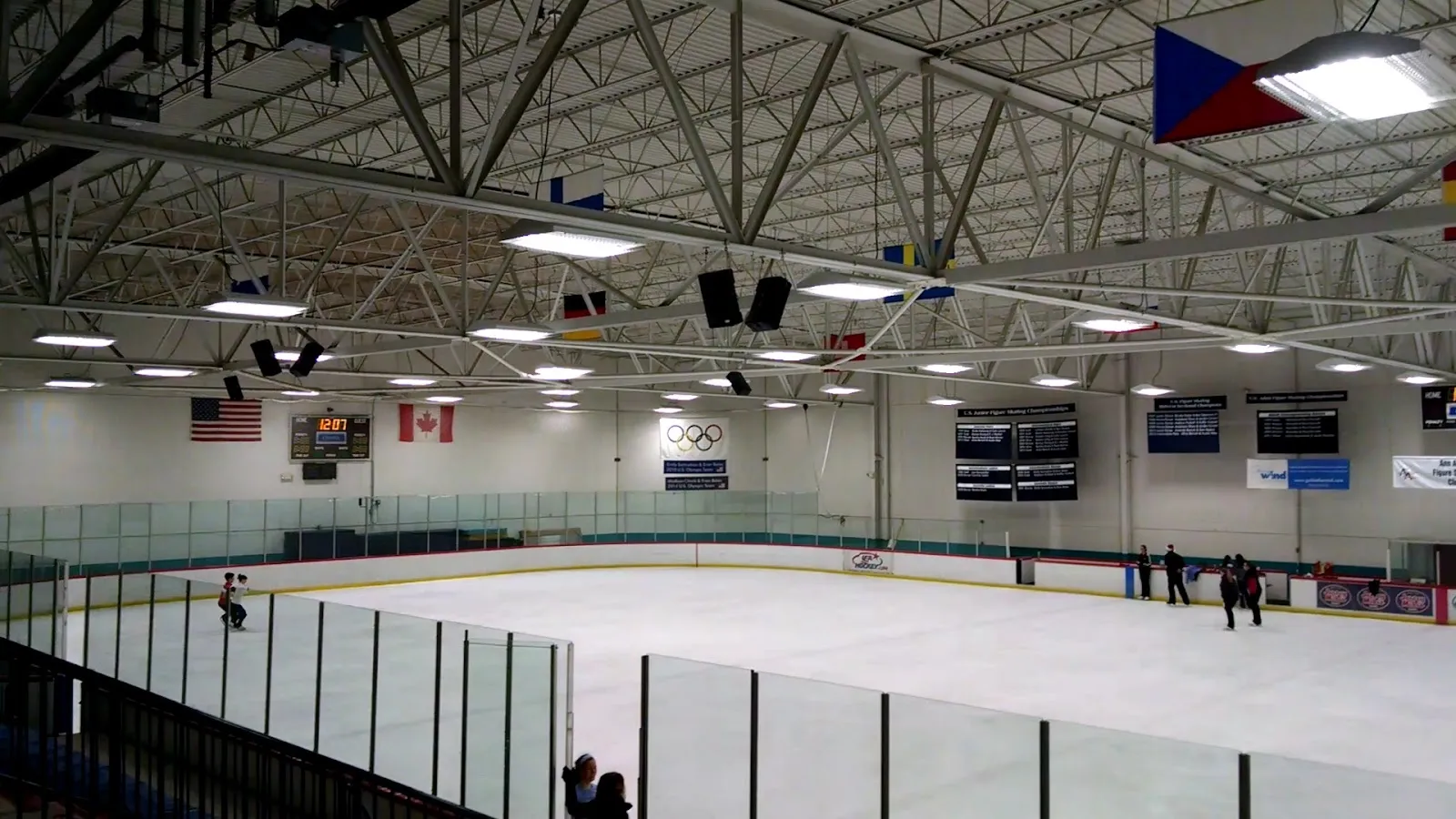 A wide empty sheet under a steel-trussed roof with flags of several nations hung in the corner and a blue scoreboard bank above the far boards. — Ann Arbor Ice Cube