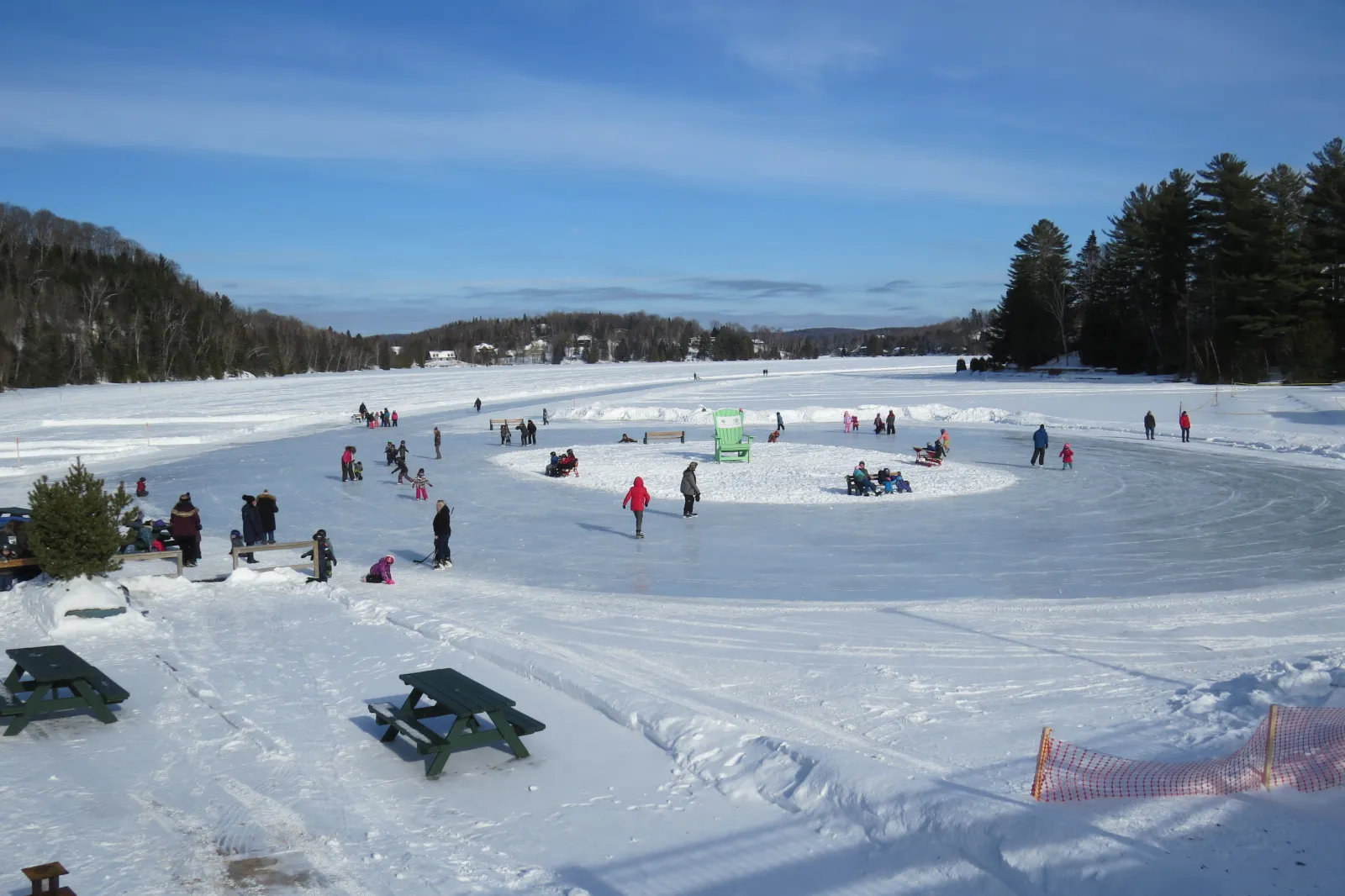 Skaters and families spread across the ice loop on Lac Masson on a clear winter day, with picnic tables on the shore and wooded hills behind. — Anneau de glace du lac Masson (Sainte-Marguerite-du-Lac-Masson)