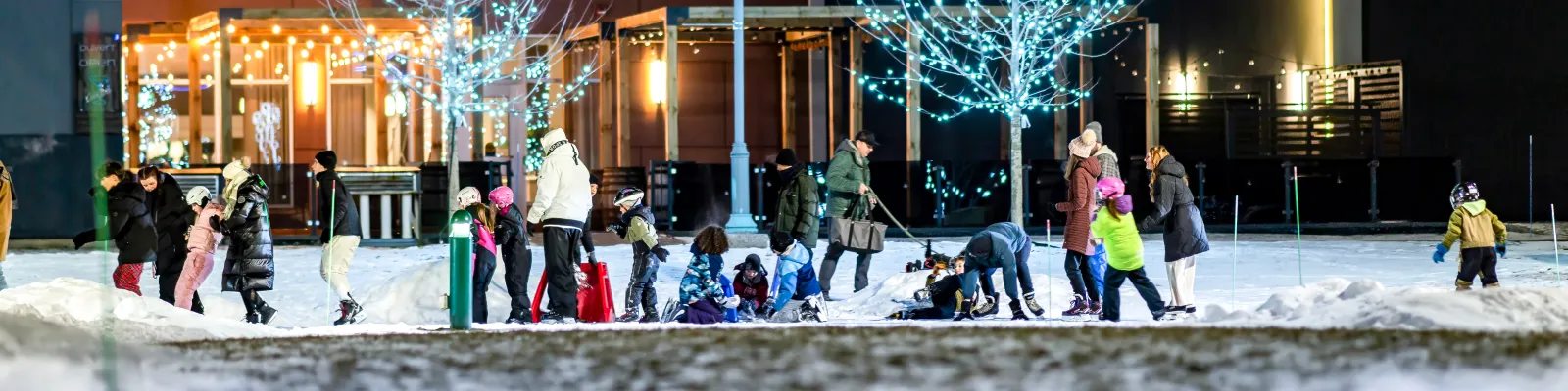 Skaters of all ages on the refrigerated ice ring at night, with the lit pavilion and illuminated trees behind. — Anneau de glace réfrigérée (Dieppe)
