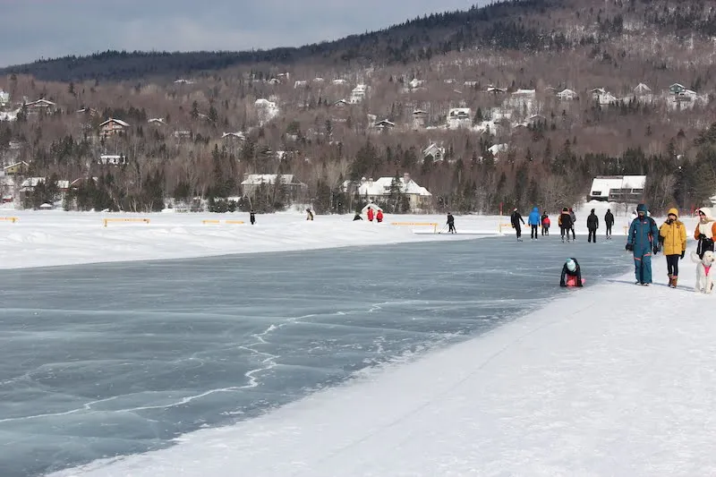 Skaters and walkers in the distance along the cleared ice of the Lac Beauport skating loop, with wooded hills behind. — Anneau de glace sur le lac Beauport (Lac-Beauport)