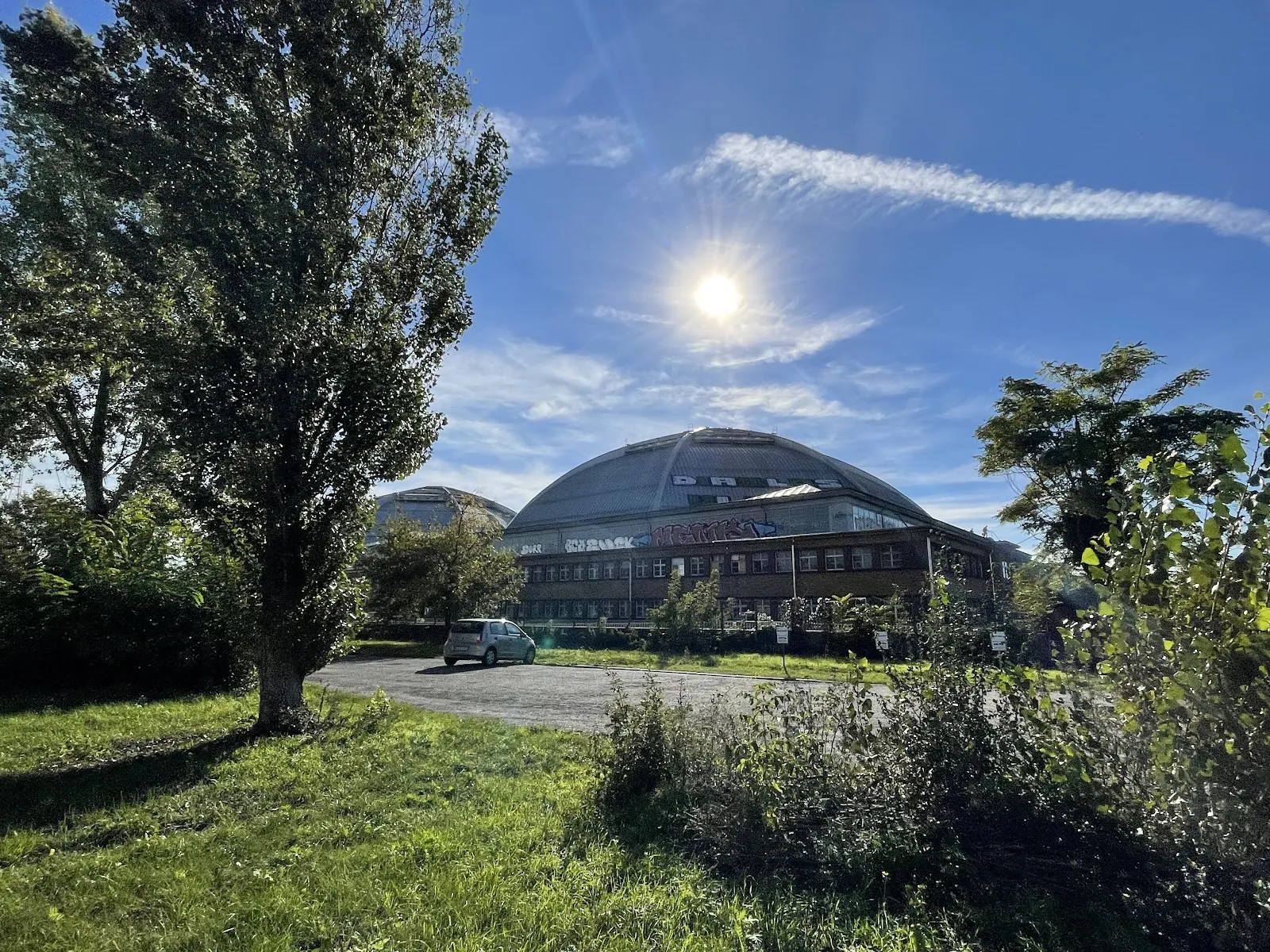 Das Kuppeldach der Arena, das hinter Bäumen und einem Schotterplatz in kräftiger Nachmittagssonne aufragt. – anona ICEDOME