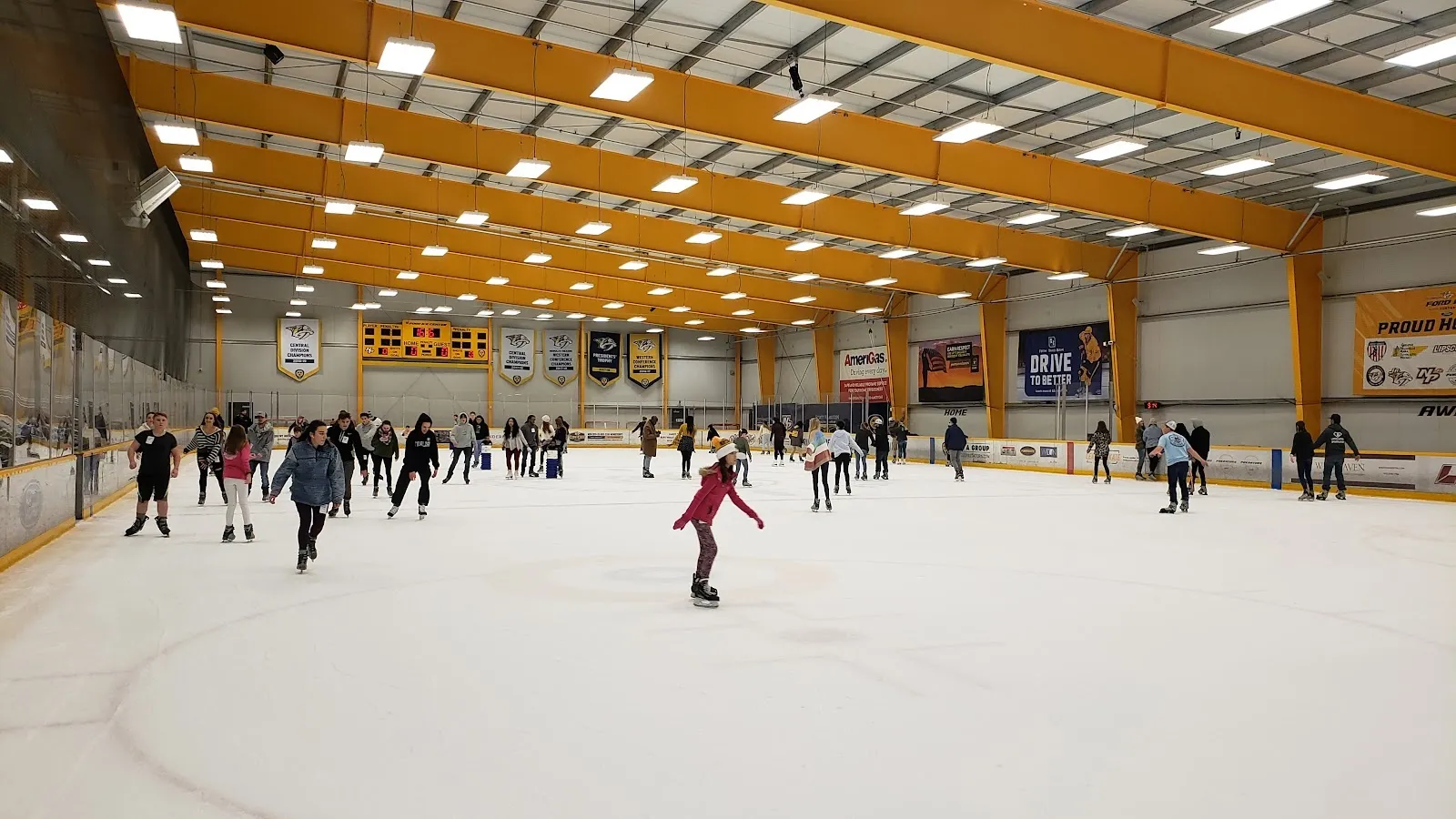 A public skating session on a wide sheet, skaters spread across the ice under a yellow-braced ceiling with Predators-coloured banners along the far boards. — Ford Ice Center – Antioch