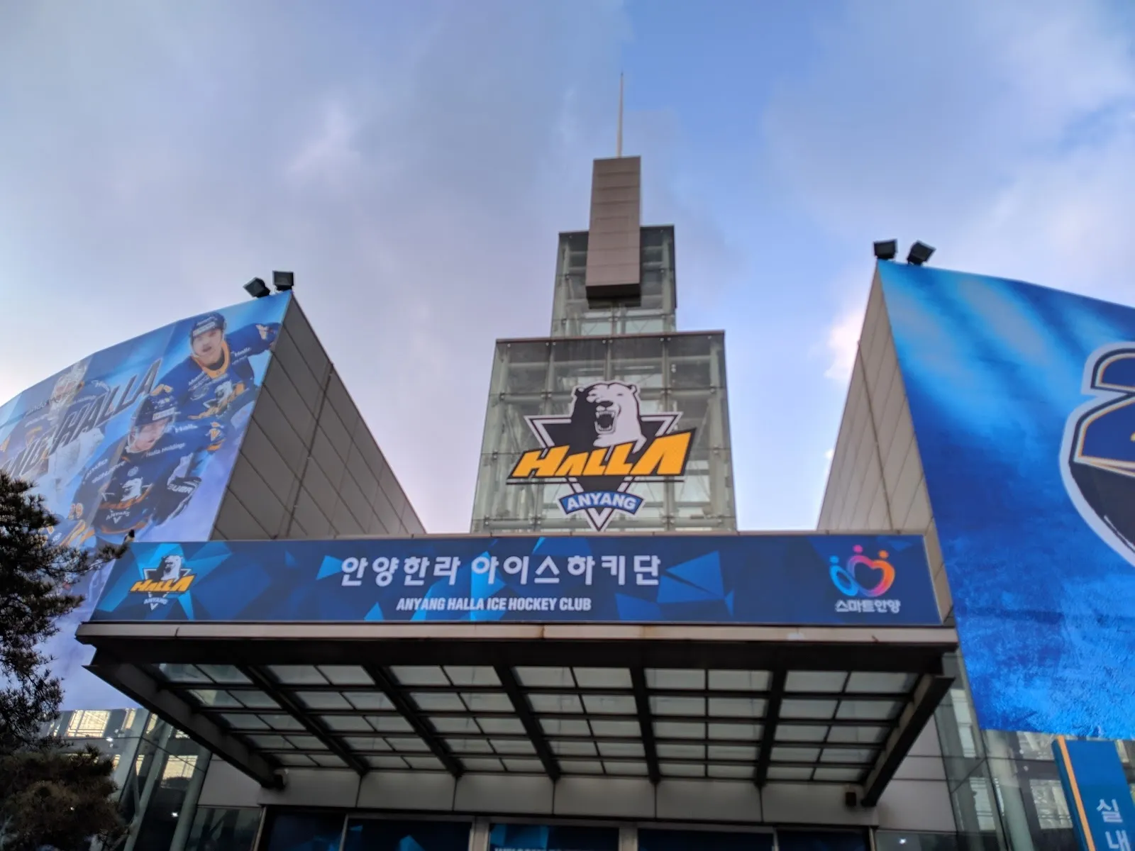 The Anyang ice arena building with its large blue HALLA banner and 25th anniversary sign on the glass front, seen from below against the sky. — Anyang Ice Arena