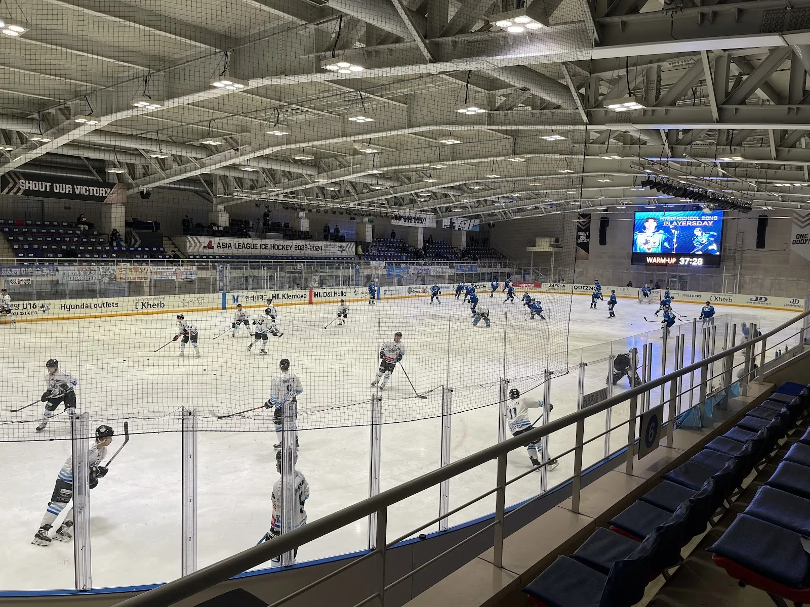 A wide view of the hall of Anyang Ice Arena from the stands during a hockey session: the steel roof trusses, the video board and the dark blue seats around the ice. — Anyang Ice Arena