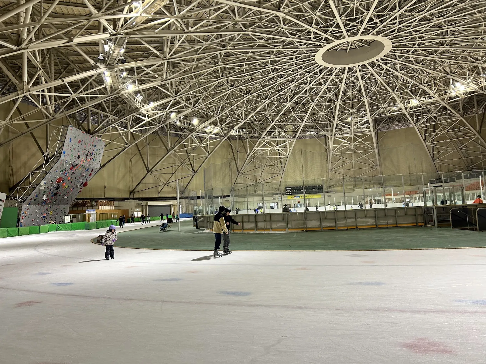 The ice inside the round steel-trussed hall of Mori Transport Arena, with a few skaters far out on the ice and a climbing wall at the left. — Aomori Prefectural Skating Rink (Mori Transport Arena)