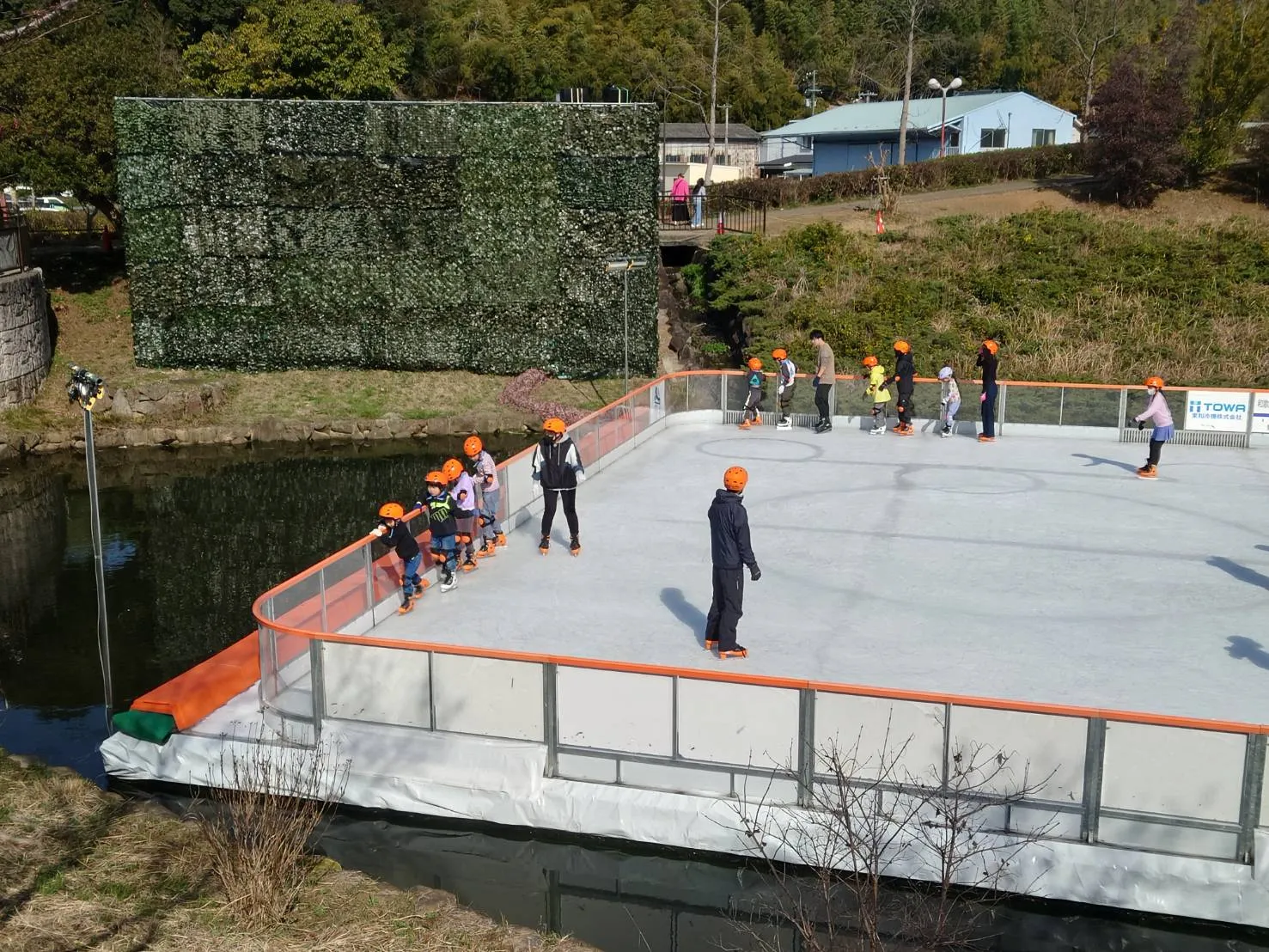 四季の郷公園の池のそばに作られた屋外の仮設リンク。オレンジ色の縁取りのフェンスがあり、オレンジのヘルメットをかぶった子どもたちが遠くで滑り、背後には木々の茂る丘が見える。 — Aozora Ice Park (Shikinosato Park)