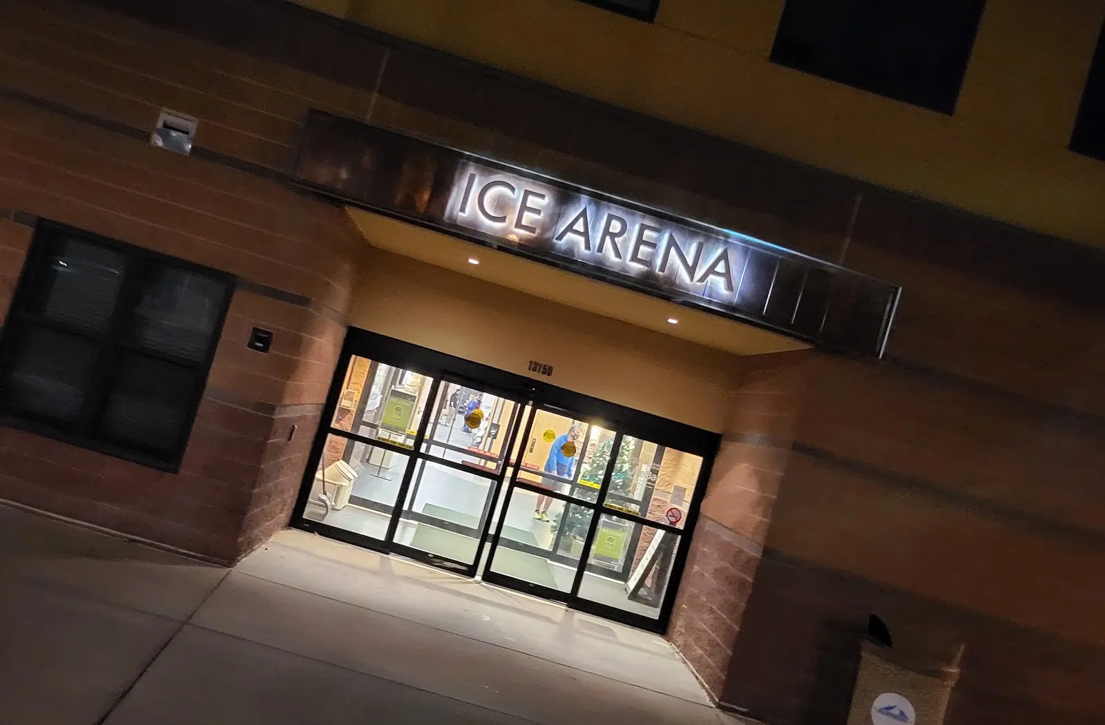 The Apex Center ice arena entrance at night: the lit ICE ARENA letters over the glass doors at 13150, in the brick and stucco wall — Apex Center Ice Arena