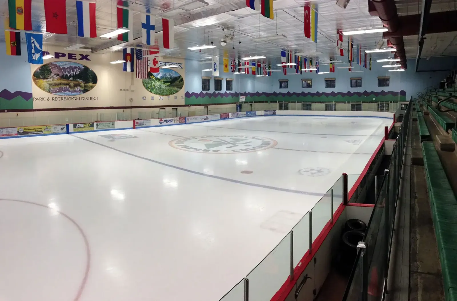 West Rink ice sheet at Apex Center, empty, with an Apex Park & Recreation District wall mural, international flags hanging from the ceiling, and green spectator seating along the boards. — Apex Center Ice Arena