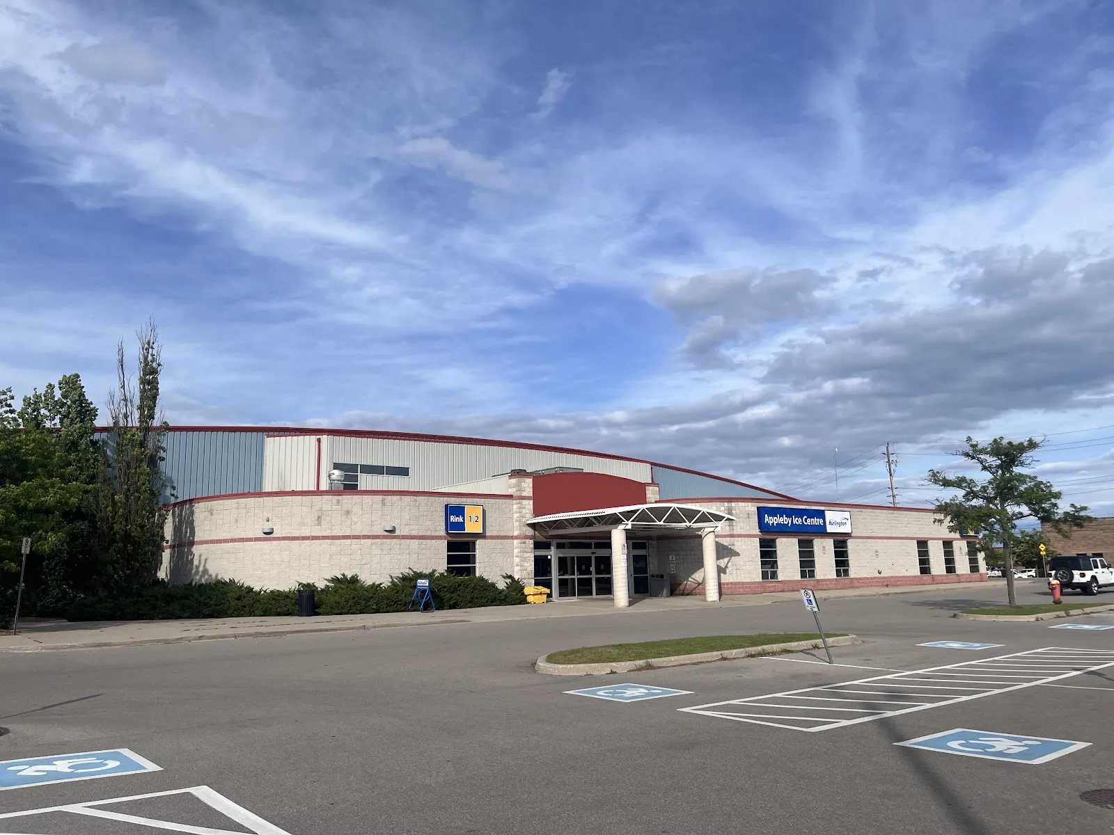Front of the low cream-and-red building with its glass entrance canopy, seen across the parking lot under a blue sky. — Appleby Ice Centre