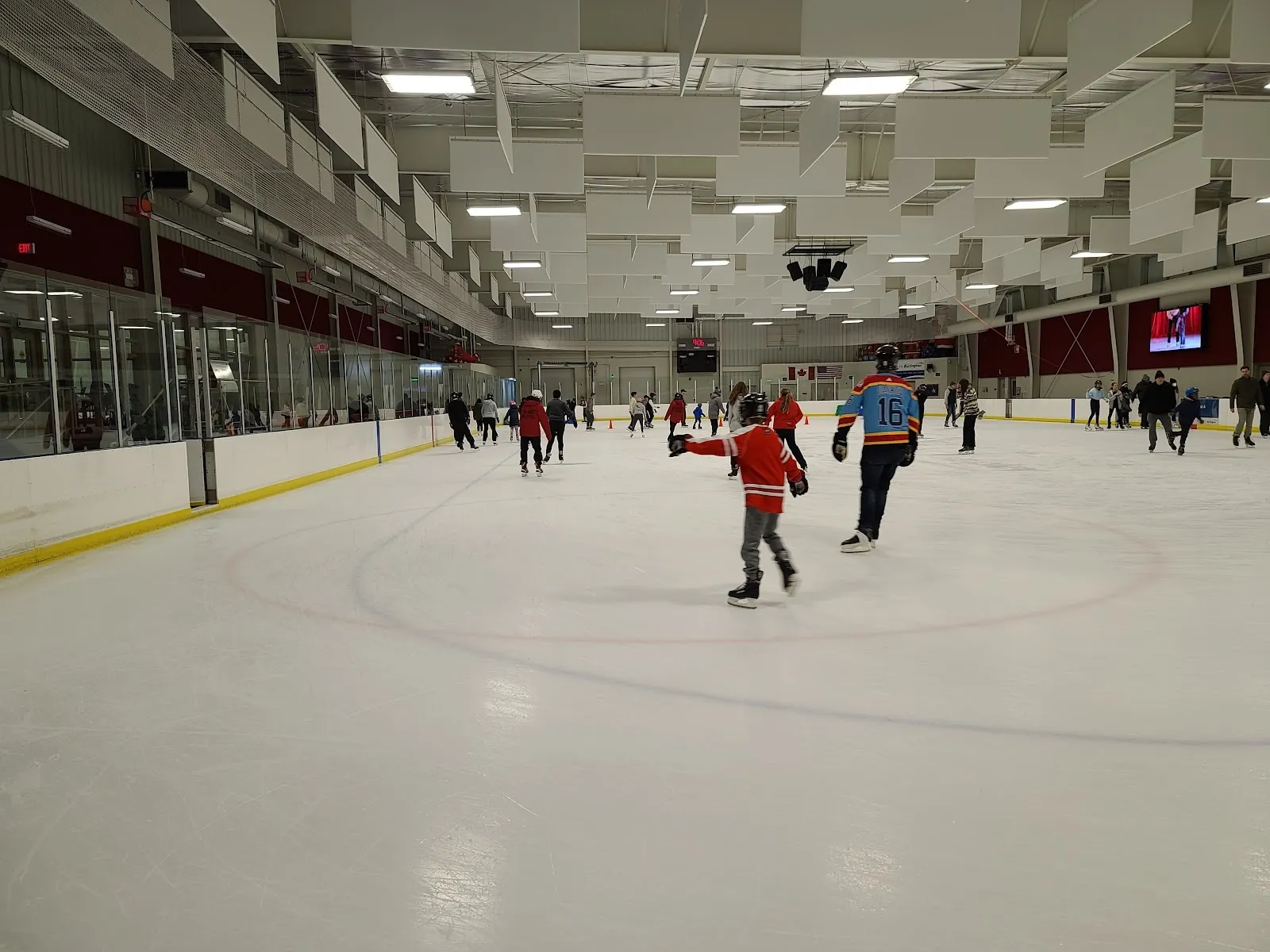 A wide view of the sheet seen from the glass, the empty white ice with a faceoff dot and a steel-trussed roof over the long far wall. — Appleby Ice Centre