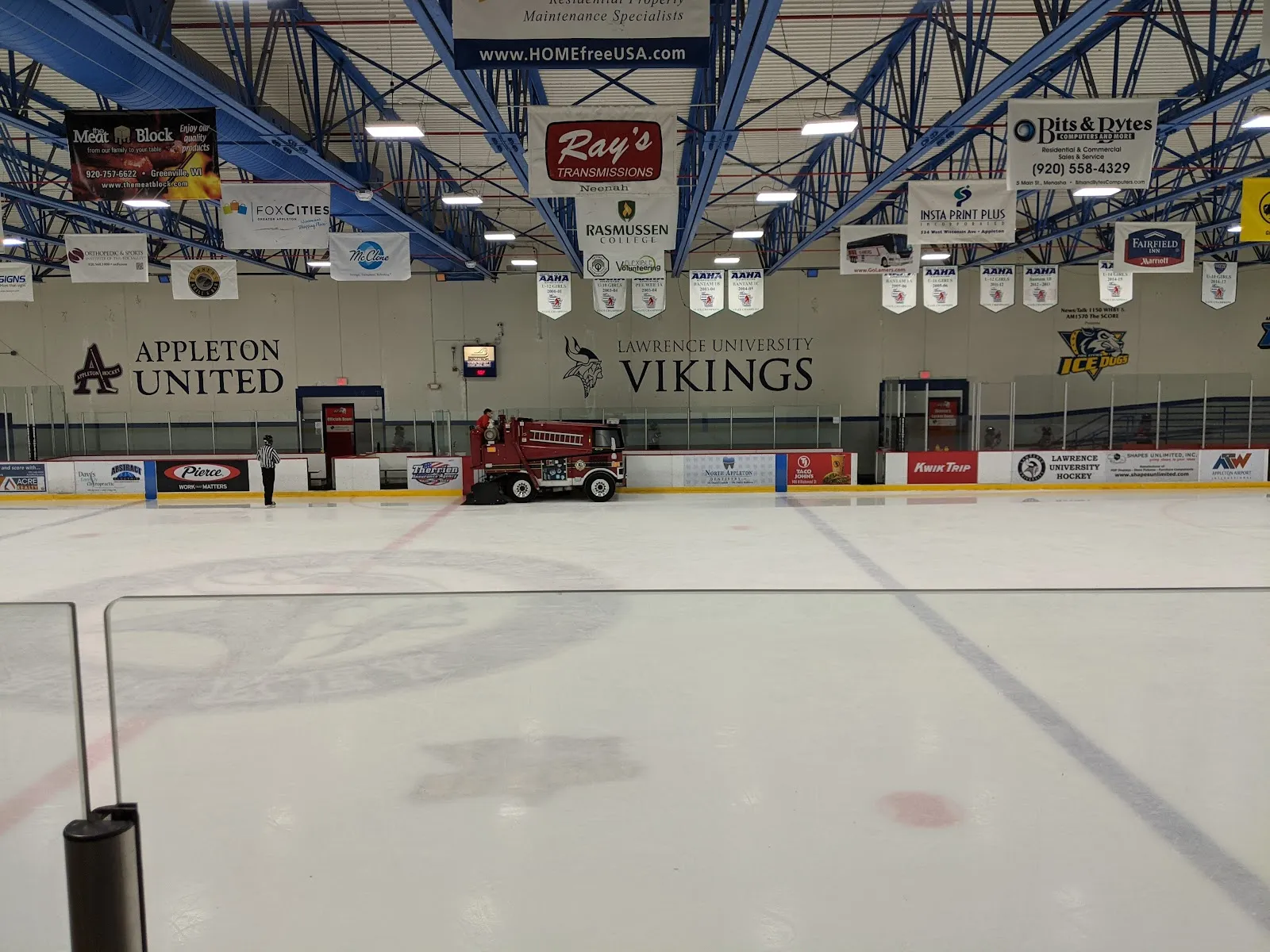 El hielo vacío desde detrás del vidrio, con las mantas de Appleton United y de los Vikings de Lawrence University y la pulidora de hielo junto a las vallas del fondo. — Appleton Family Ice Center