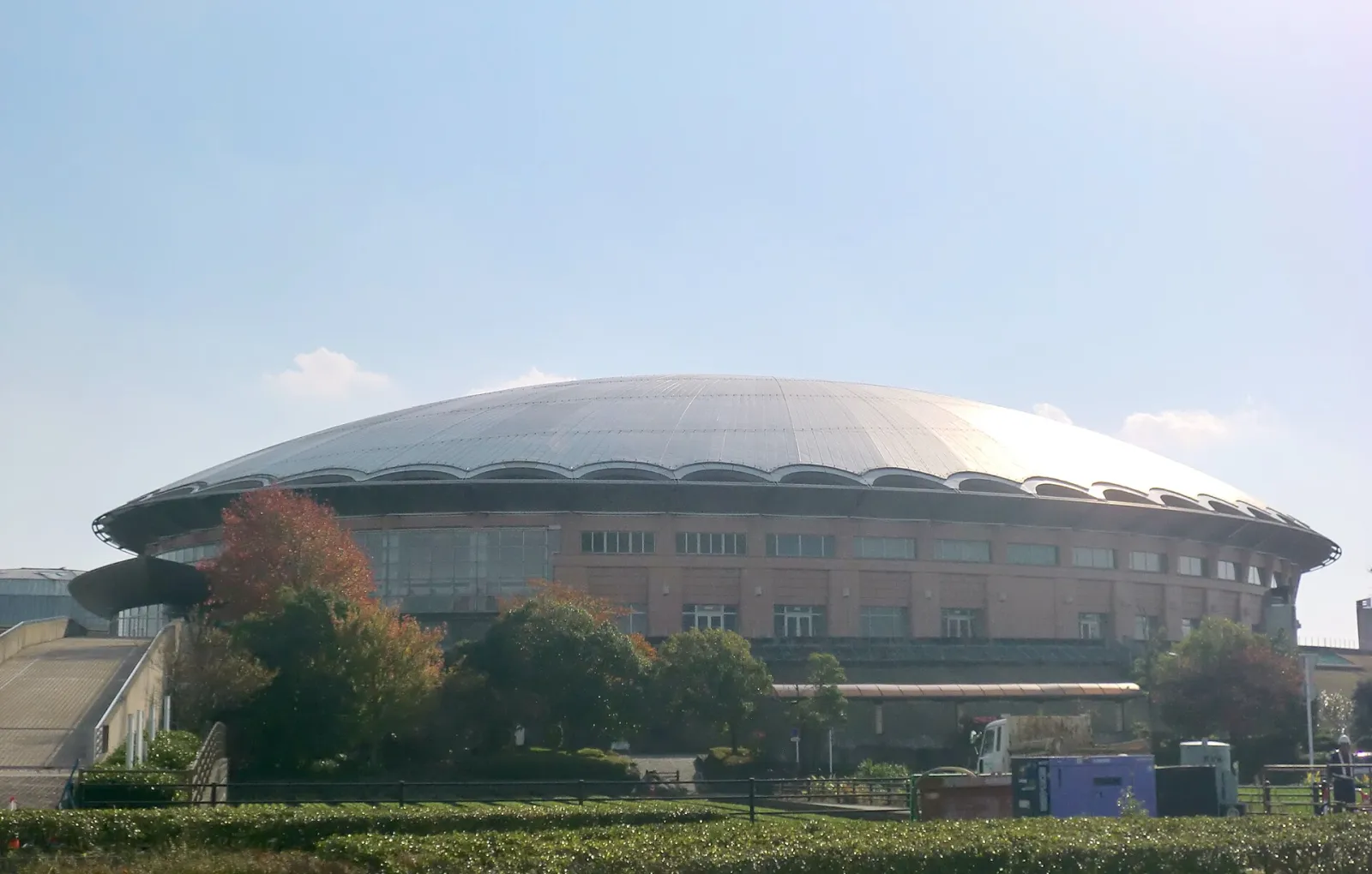 The silver dome roof of Aqua Dome Kumamoto above its brick-faced hall, seen across the park grounds in autumn. — Aqua Dome Kumamoto Ice Rink
