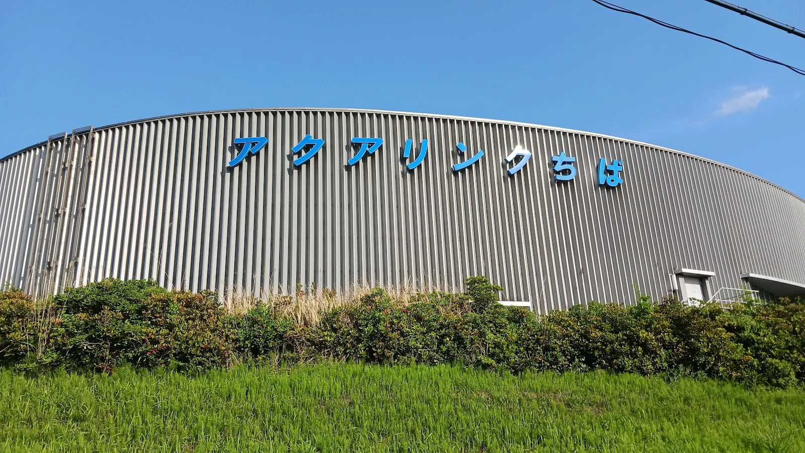 The curved ribbed-metal wall of Aqua Rink Chiba with its blue katakana name sign, seen from the grass outside under a blue sky. — Aqua Rink Chiba