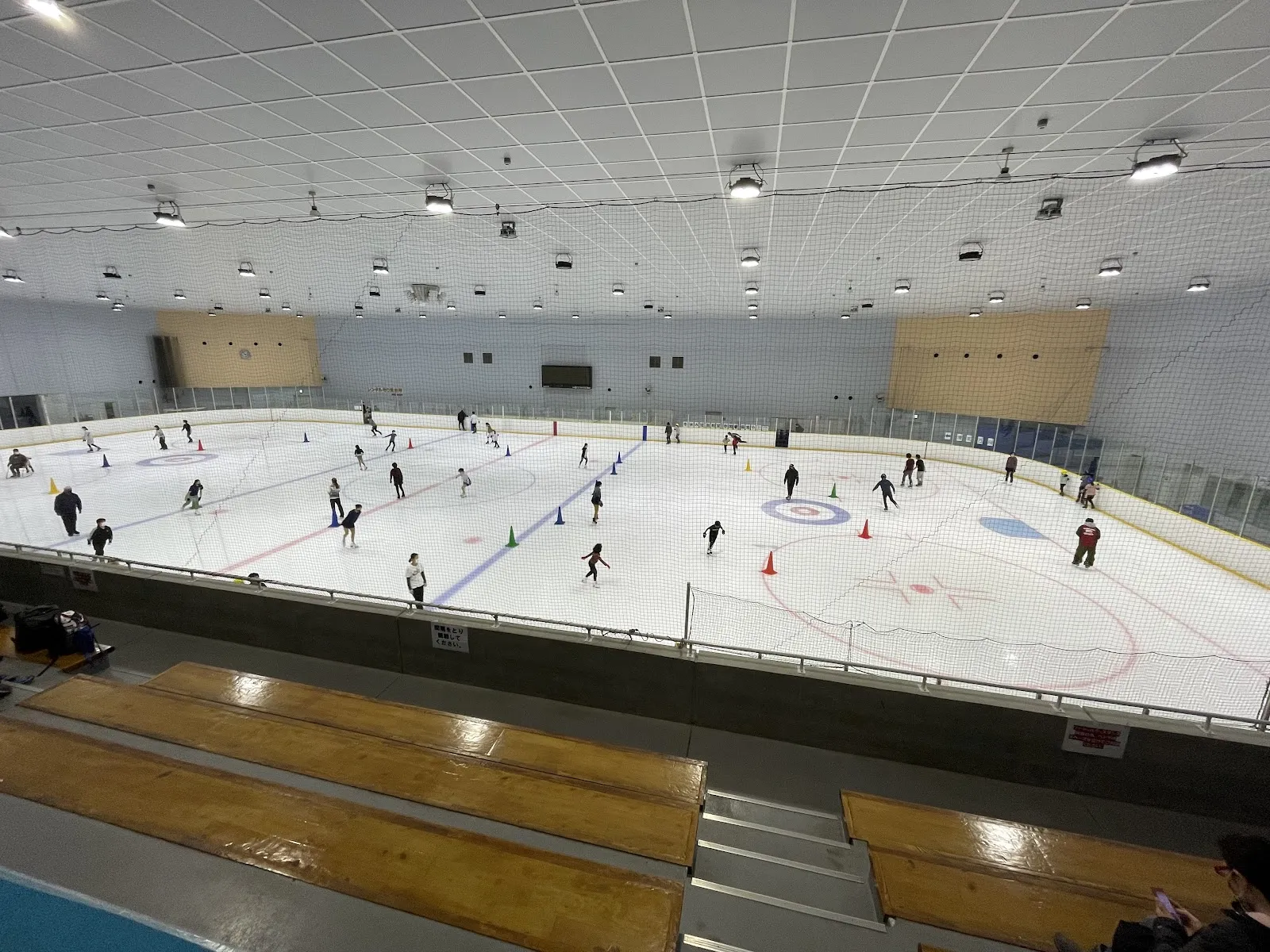 The wide ice sheet of Aqua Rink Chiba seen from the wooden stands, with skaters spread across the surface under the white ceiling. — Aqua Rink Chiba
