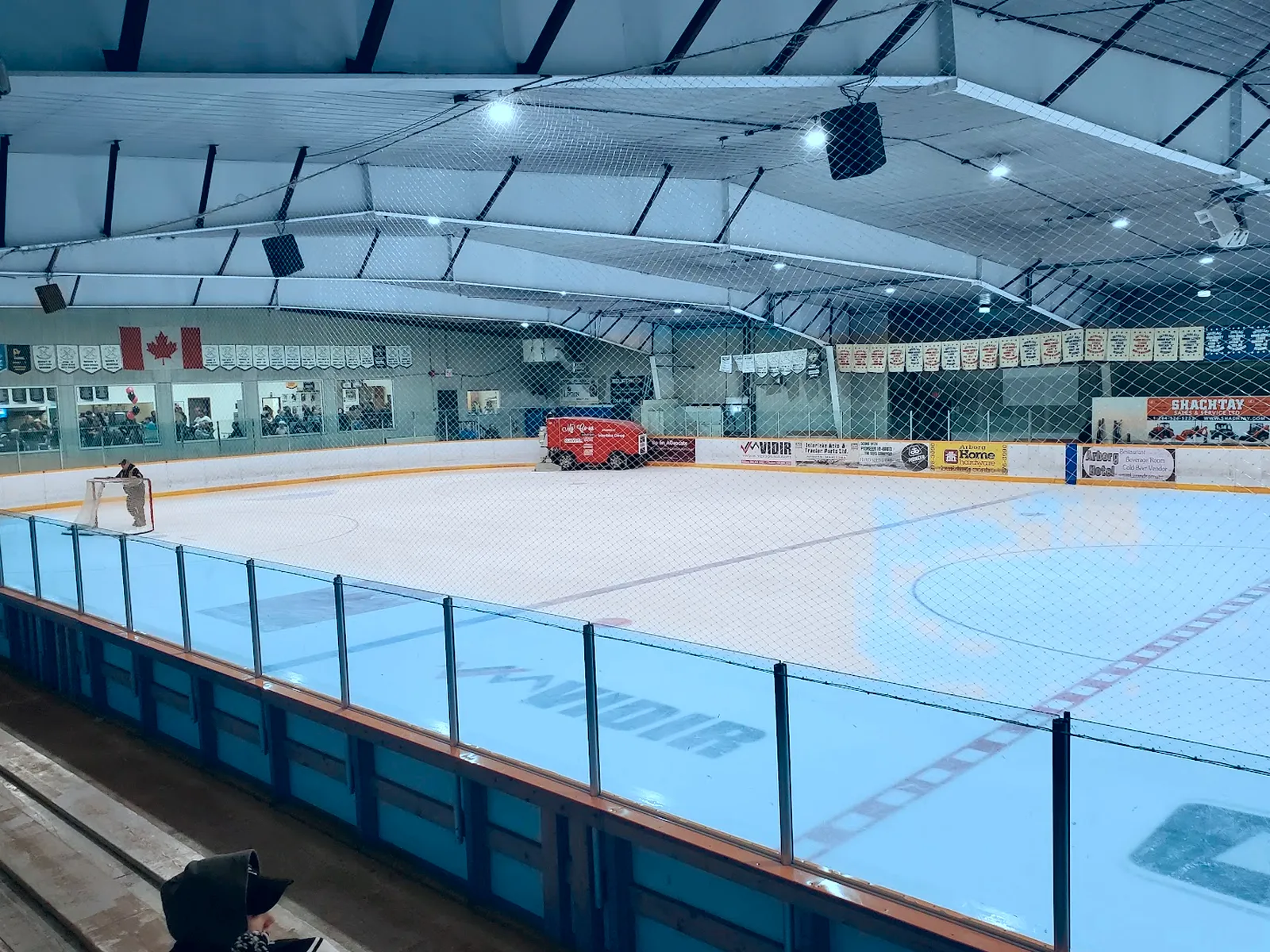 El hielo vacío de Arborg desde detrás del vidrio, con la pulidora de hielo junto a las vallas del fondo y mantas y banderas a lo largo del muro del extremo — Arborg & District Arena