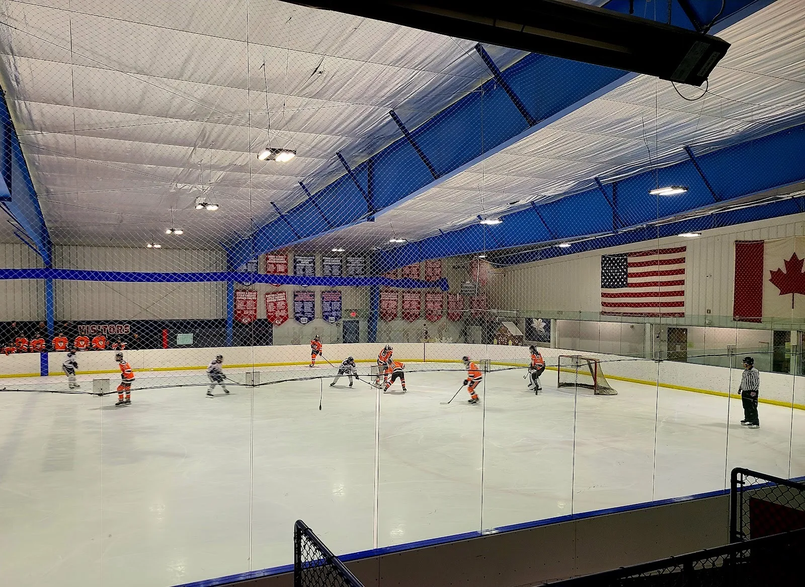 Una pista de Arctic Edge of Canton desde detrás del vidrio, con vigas azules en el techo, mantas de campeonatos, las banderas de Estados Unidos y Canadá y un partido juvenil — Arctic Edge Ice Arena (Canton)