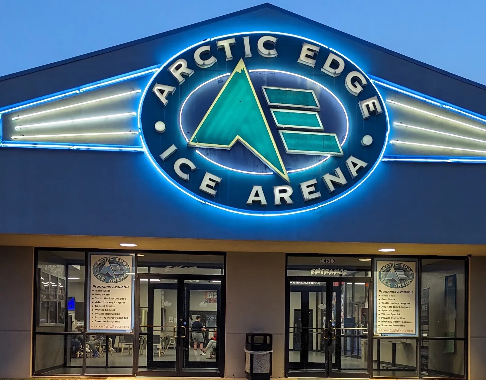 La entrada del Arctic Edge Ice Arena al atardecer, con su letrero redondo iluminado de azul sobre dos juegos de puertas de vidrio — Arctic Edge Ice Arena