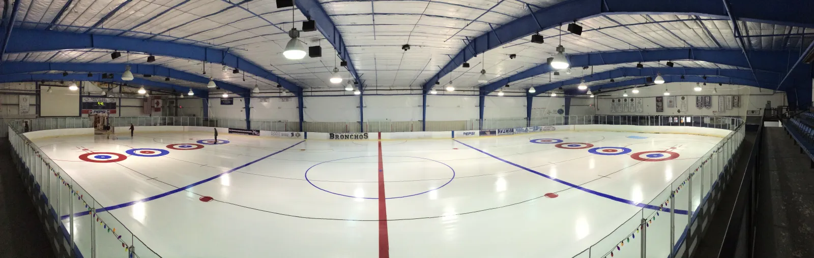 Amplia panorámica interior de una pista de hielo vacía, con las armaduras azules del techo, las letras "BRONCHOS" en las vallas, mantas de campeonato y círculos tipo diana de curling pintados en el hielo en cada extremo. — Arctic Edge Ice Arena
