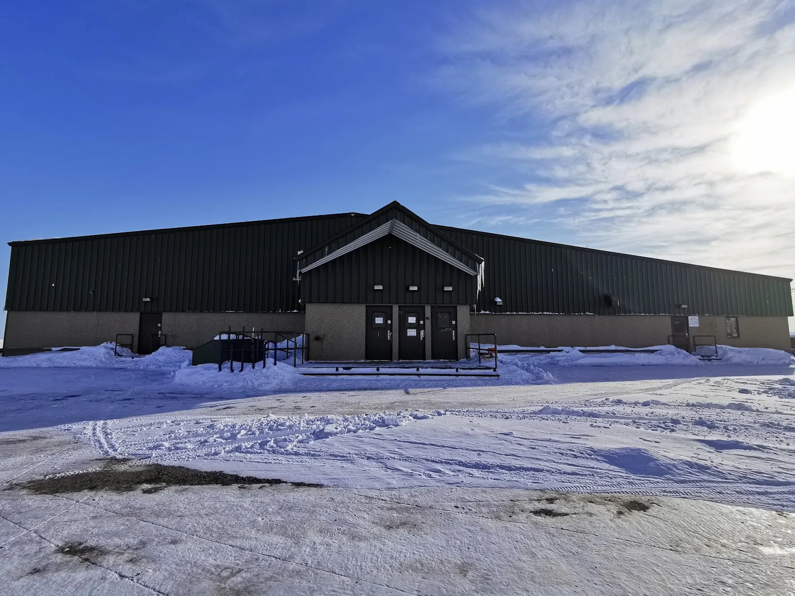 The low dark Arctic Winter Games Arena building in Iqaluit with a snow-covered lot in front and a pale sky. — Arctic Winter Games Arena