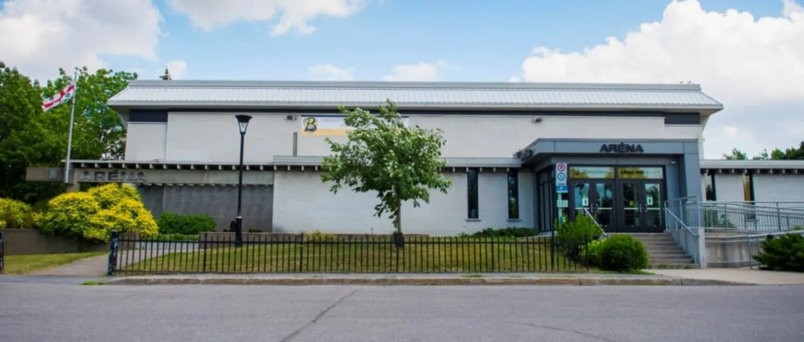 White brick single-storey arena building seen from the street, with an illuminated 'ARENA' sign over the glass entrance and a flag pole out front. — Aréna Ahuntsic