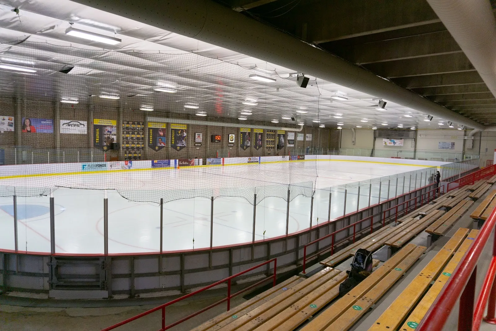 Aréna Ahuntsic's empty sheet from the wooden bleachers, with the Braves banners on the brick wall and the netting along the boards — Aréna Ahuntsic