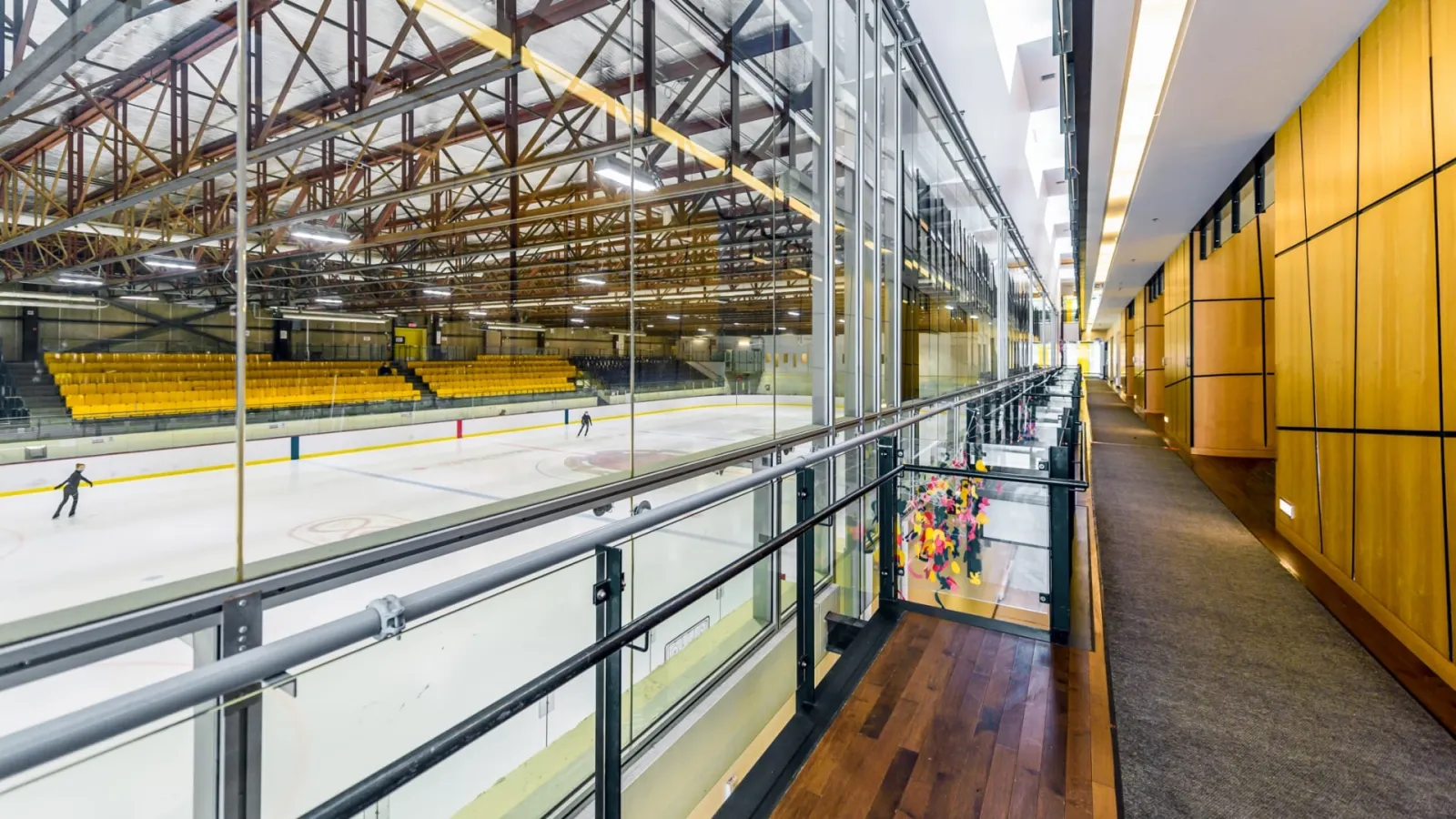 Full ice sheet seen from an upper glass-walled walkway, with yellow spectator seating along the boards and two skaters far below on the ice. — Aréna André-Laperrière