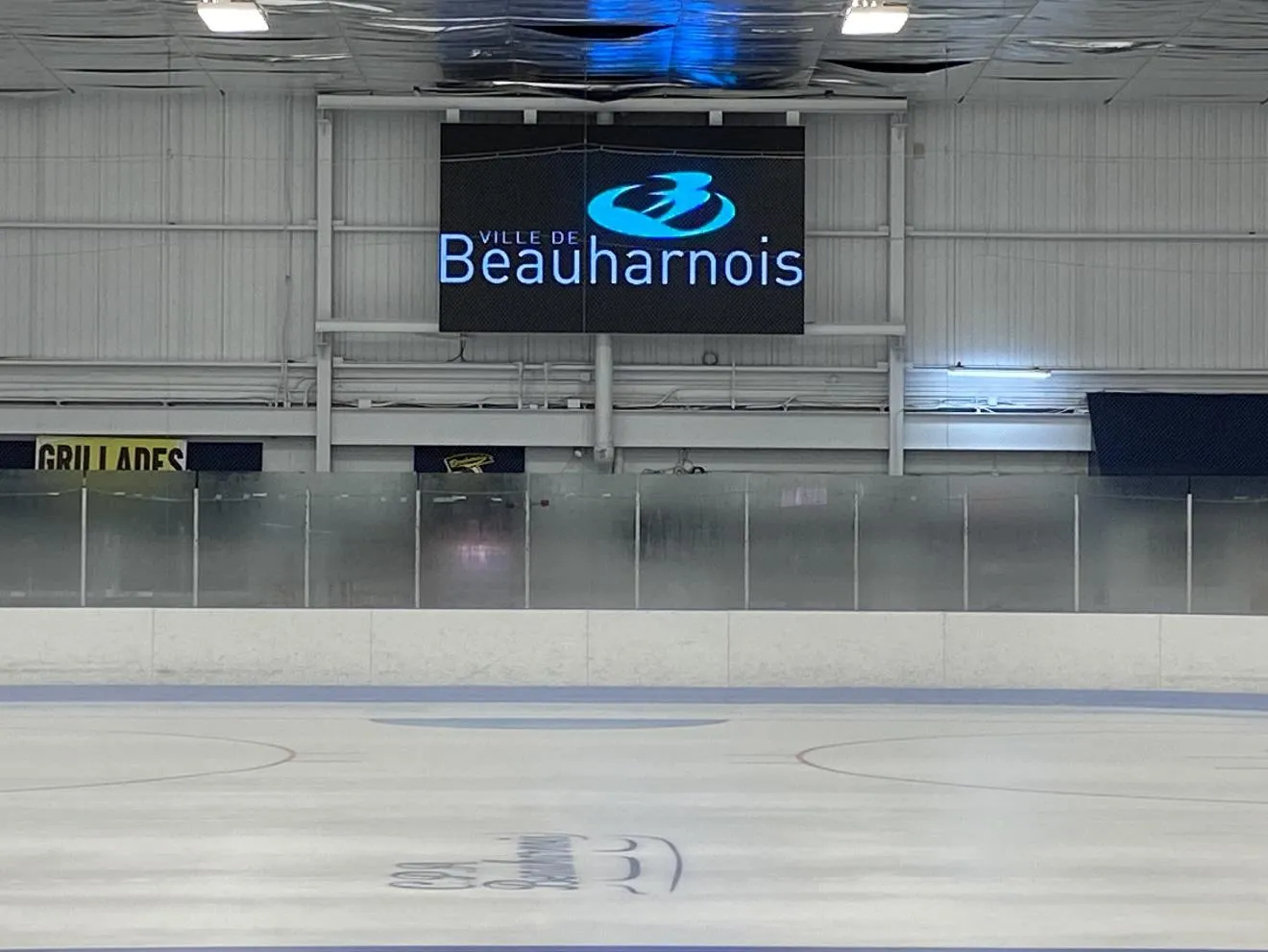 The arena ice with the Beauharnois logo painted at centre, the boards and glass, and a video screen showing the Ville de Beauharnois logo above the far side. — Aréna André-Richard