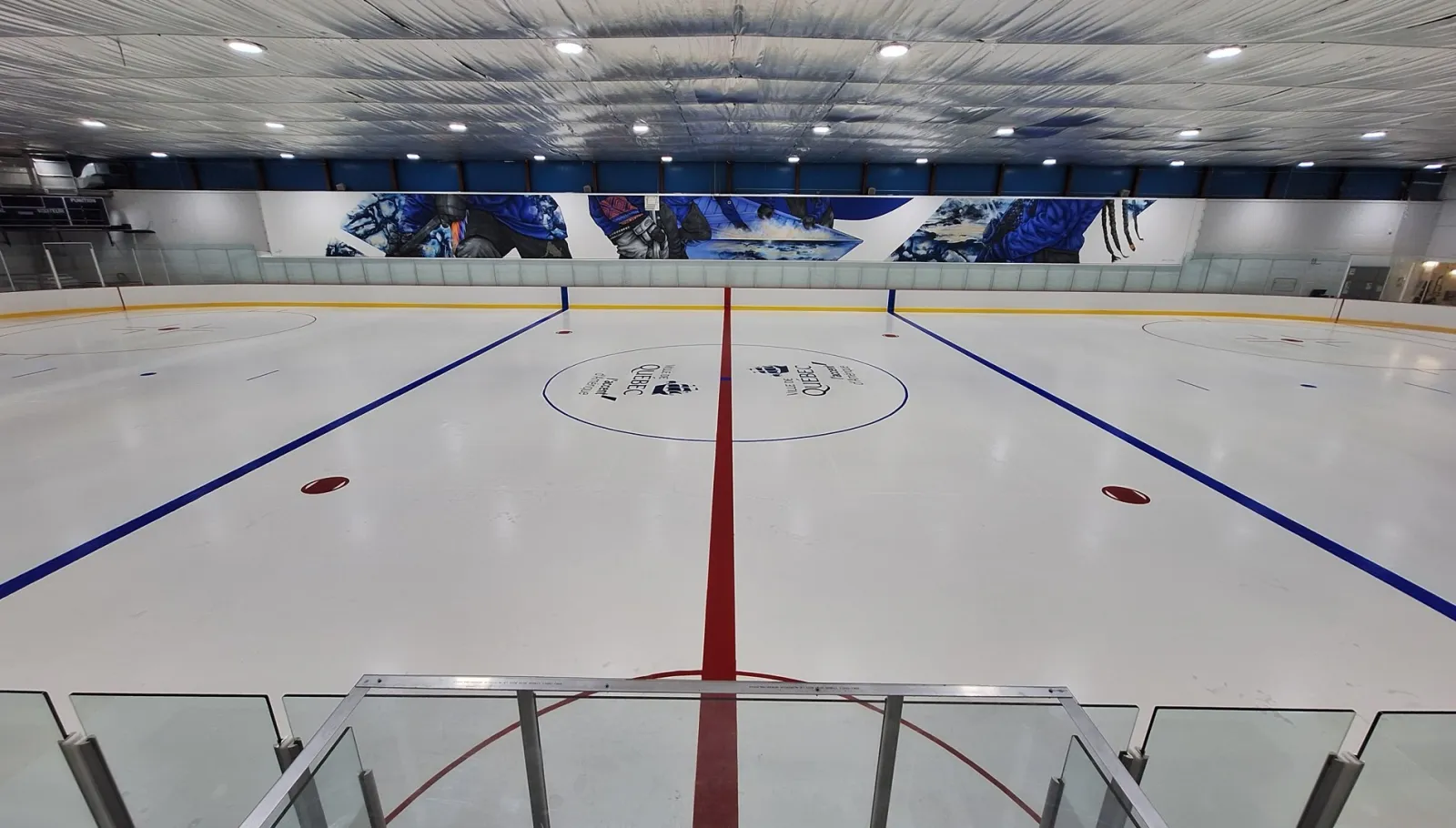 La glace vide vue du centre, derrière la baie vitrée, avec une murale sur le thème du hockey le long du mur du fond et un plafond blanc garni de rangées de lumières. — Aréna Bardy