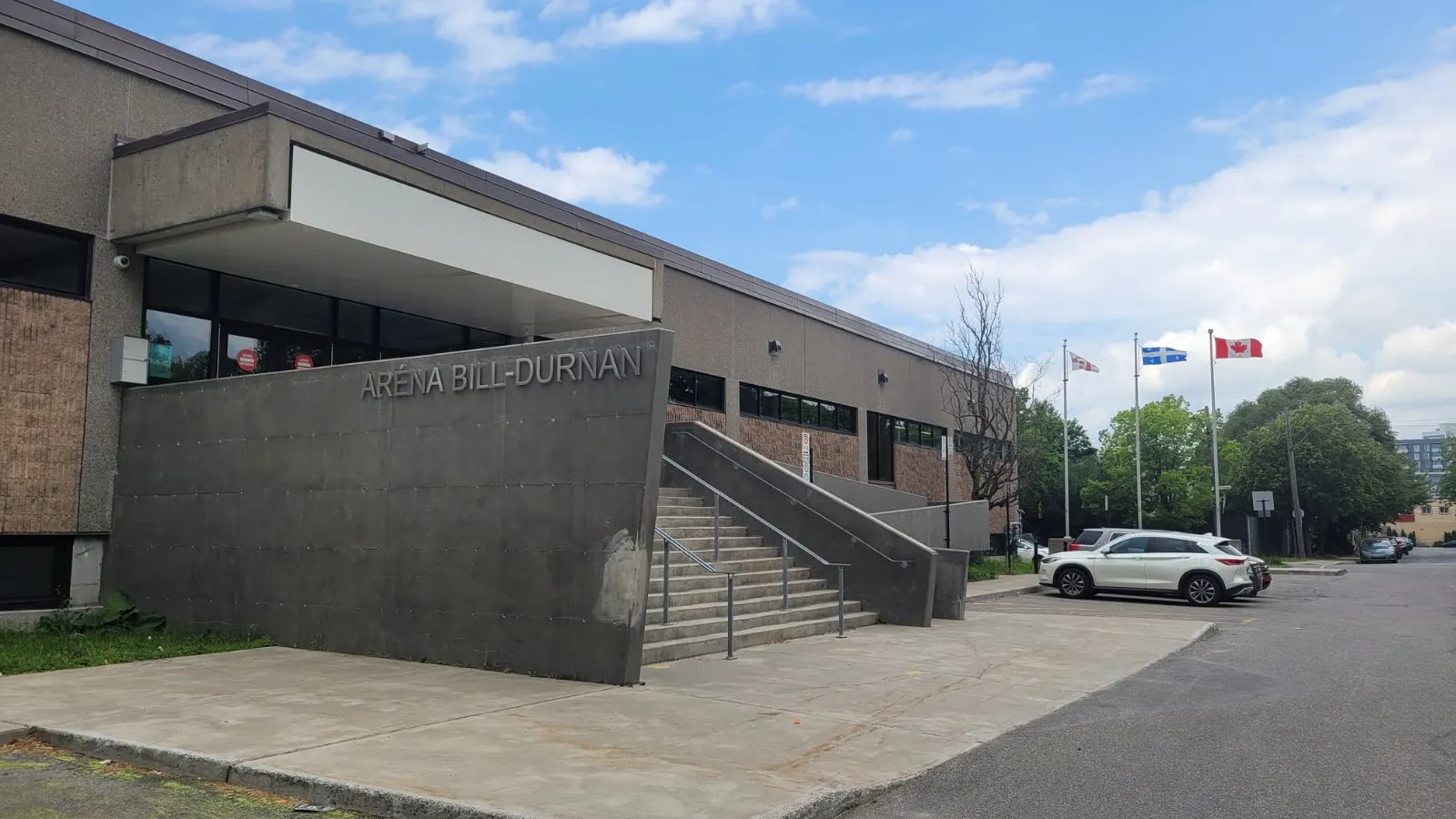 Front entrance of Aréna Bill-Durnan, concrete facade with raised "ARÉNA BILL-DURNAN" lettering, stairs to the door, and a parking lot with Quebec/Canada flags in the background. — Aréna Bill-Durnan