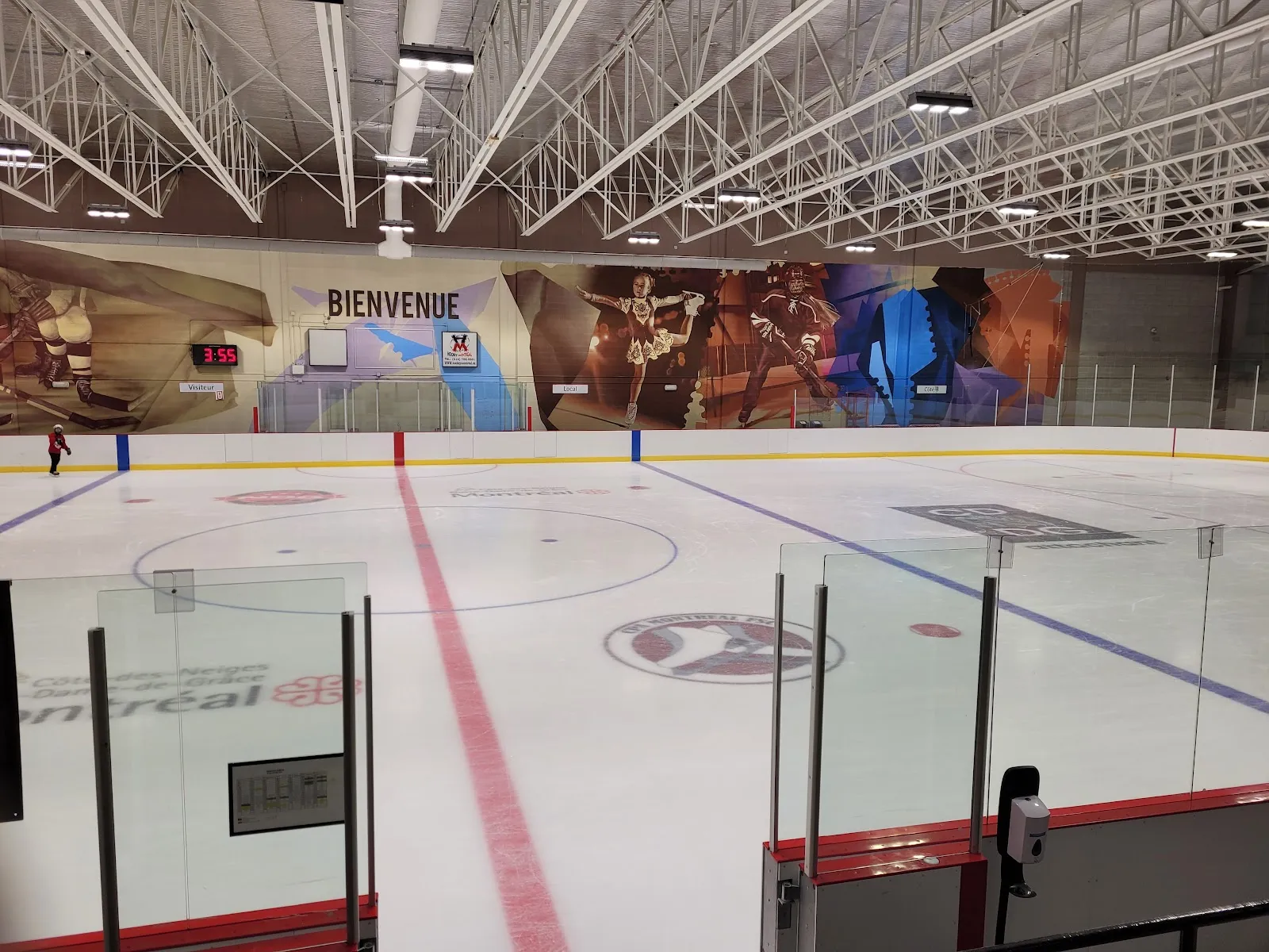 Full ice sheet from the stands, red/blue lines and a Côte-des-Neiges–Notre-Dame-de-Grâce/Montréal borough logo on the ice, a "BIENVENUE" mural on the far wall, and one skater in the distance. — Aréna Bill-Durnan