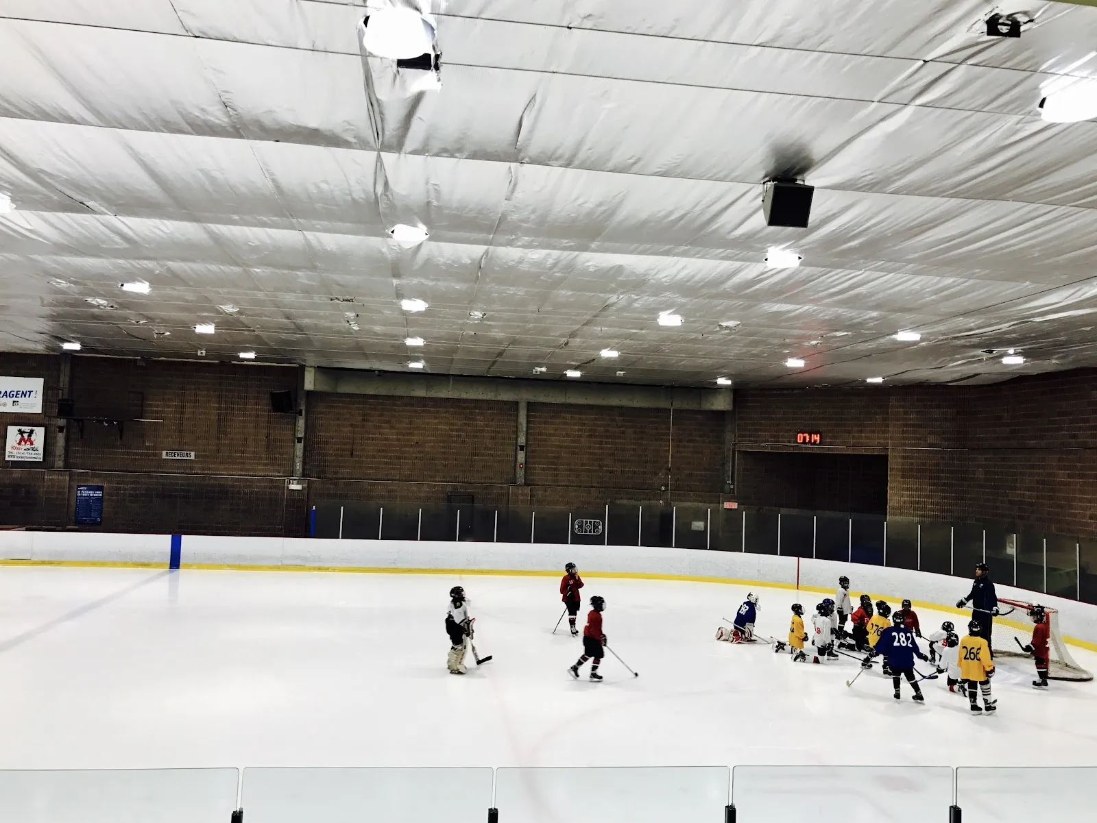 Un entraînement sur une glace blanche vu des gradins, des joueurs rassemblés au centre sous un plafond blanc plissé, avec un mur de briques derrière. — Aréna Camillien-Houde