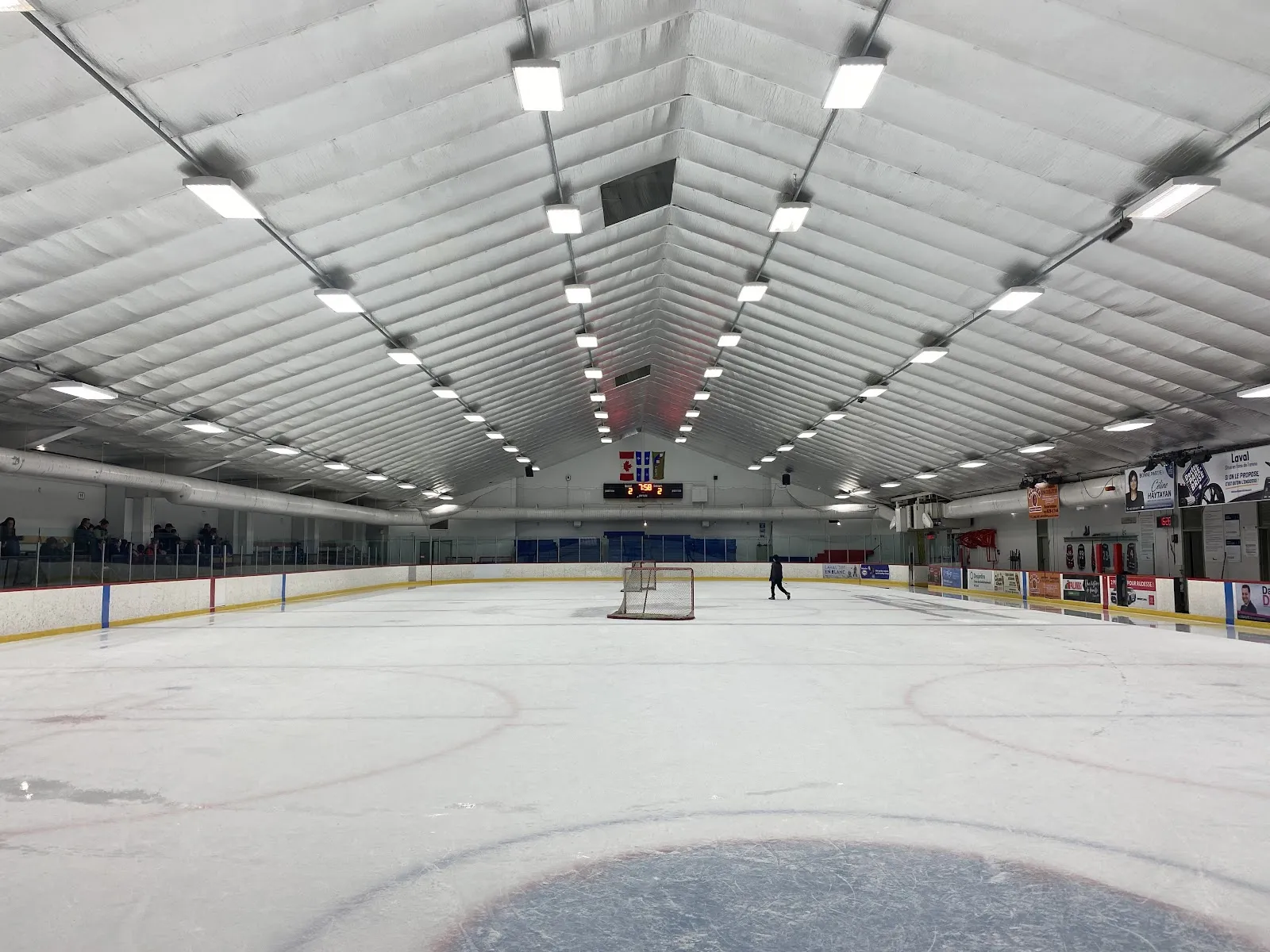 The ice sheet seen from the boards under the long insulated roof, with a goal and a single skater far down the rink, the boards and sponsor panels along the far side. — Aréna Cartier (Laval)