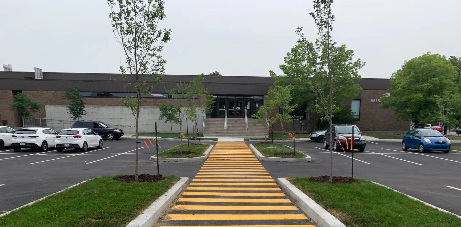 La entrada principal de Aréna Chaumont vista desde el otro lado de un estacionamiento, un edificio bajo de ladrillo café con un paso peatonal rayado que lleva a las puertas de vidrio y un letrero de "AREN..." parcialmente visible en la pared. — Aréna Chaumont