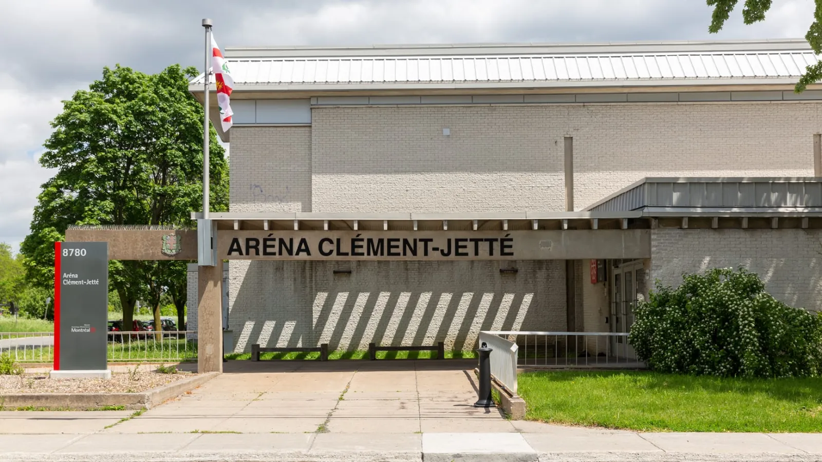 La marquesina techada de la entrada de Aréna Clément-Jetté con las letras en relieve "ARÉNA CLÉMENT-JETTÉ", una bandera de Montréal y el pilón con la dirección al frente. — Aréna Clément-Jetté