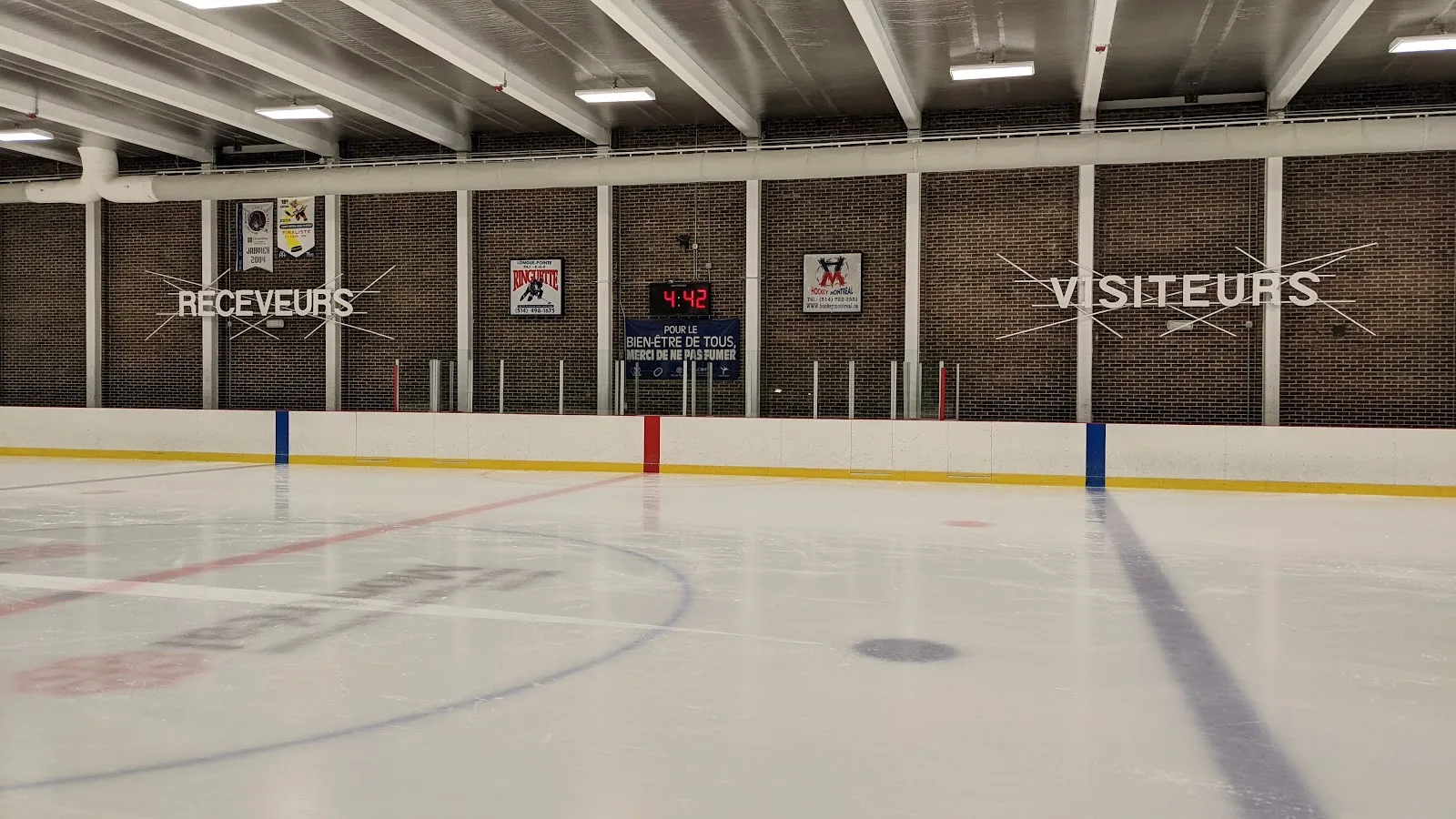 Pista de hielo vacía hacia las vallas del fondo, con letreros de las bancas "RÉCEVEURS" y "VISITEURS", un reloj de juego y una manta de Hockey Montréal en el muro de ladrillo; no hay personas visibles. — Aréna Clément-Jetté
