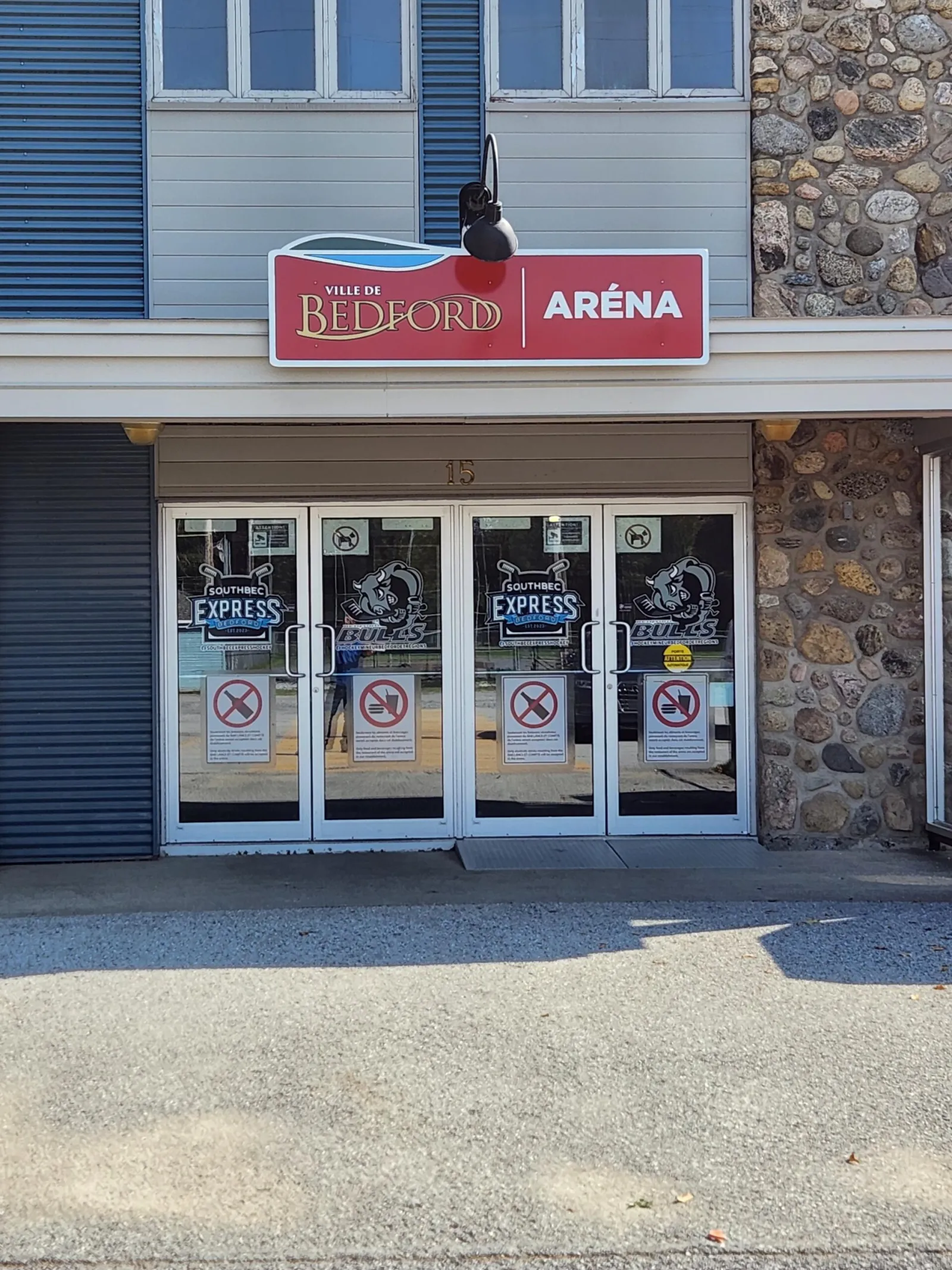The entrance of the Bedford arena under the red Ville de Bedford Aréna sign, with glass doors carrying Southbec Express hockey decals and stone facing on the wall. — Aréna de Bedford