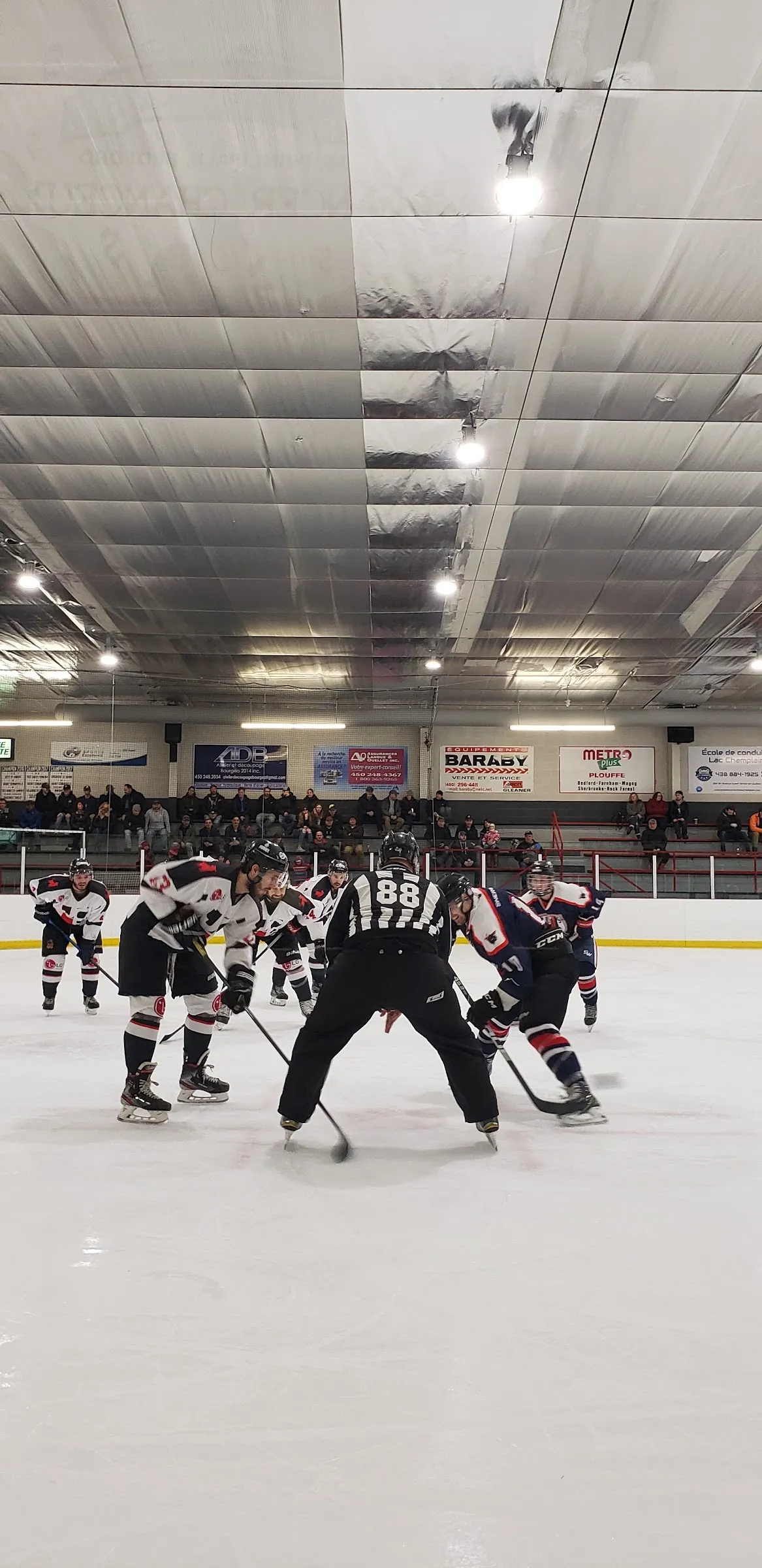 A faceoff at centre ice in a portrait view, players and an official spread across the white ice with a bright ceiling and stands behind. — Aréna de Bedford
